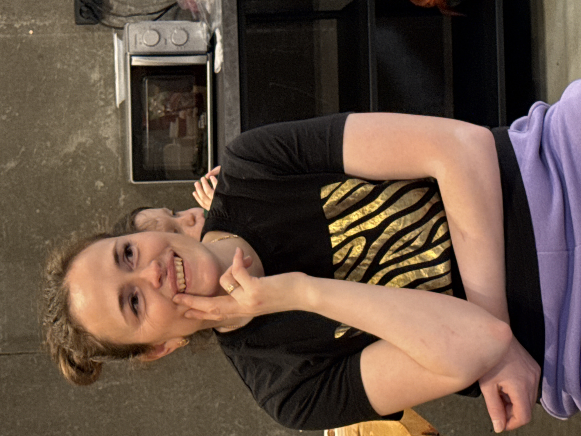 A woman with light skin, light brown hair in a low bun, wearing a black t-shirt with gold zebra stripes and a gold necklace, is smiling and touching her chin with her right hand. She has a wedding ring on her left hand. A child with light skin is partially visible behind her. They are positioned in front of a black cabinet with shelves and a microwave oven on a gray wall. No information in the image confirms a location in Yekaterinburg, Russia.
