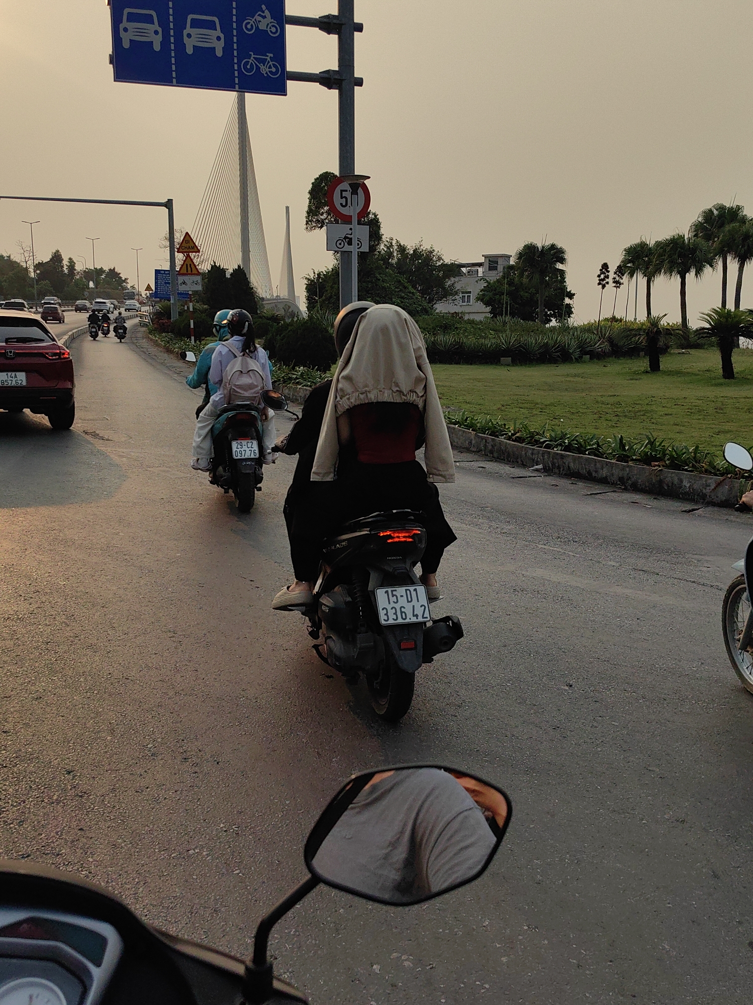 The image depicts a street scene in Hong Gai Ward, Vietnam, during daylight. Several people are riding motorbikes on a paved road. A car is visible in the background. Traffic signs indicate lanes for cars, motorbikes, and bicycles. A bridge stands in the distance. The sky is overcast, and palm trees are on the right side of the road. The image is taken from the perspective of someone riding a motorbike, as part of the vehicle, including a mirror, is visible in the foreground. License plates are visible on some of the motorbikes.
