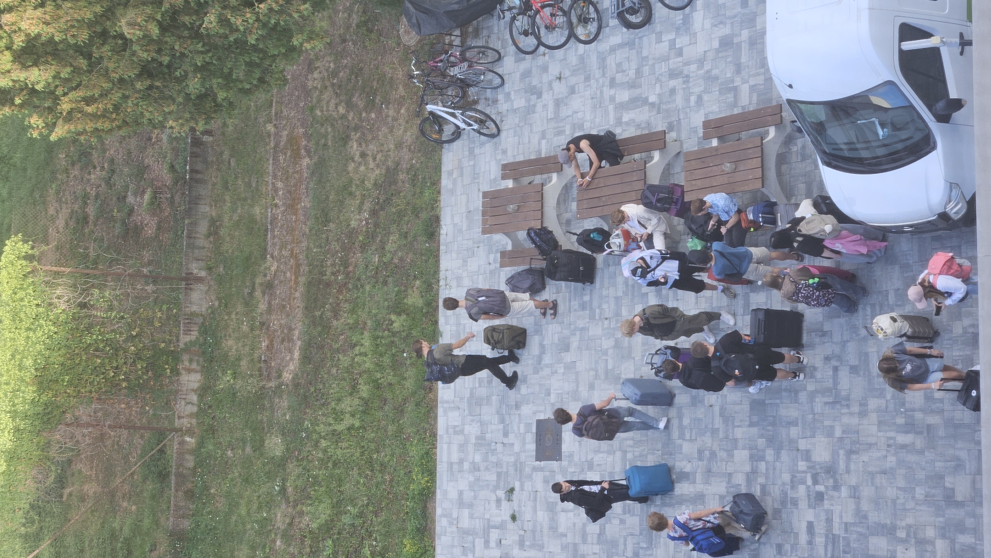 This is an overhead, vertical, full-shot photograph taken outdoors during daylight. The scene depicts a group of people with luggage on a paved area adjacent to a grassy embankment with a fence. The pavement is composed of rectangular gray tiles.
In the upper left, a grassy hill slopes upwards, covered with various vegetation and a chain-link fence.
In the mid-ground, several individuals are scattered across the paved area. Some are standing, others are sitting at picnic tables. Luggage, including suitcases and backpacks, is visible throughout the scene. Bicycles are parked on the right side.
In the foreground, the front portion of a white van is visible on the right.
The people are dressed in casual clothing, and their activities suggest they are either arriving or departing, possibly from a facility or event. The overall atmosphere is one of transit and waiting. The location is identified as Prešov, Slovakia.