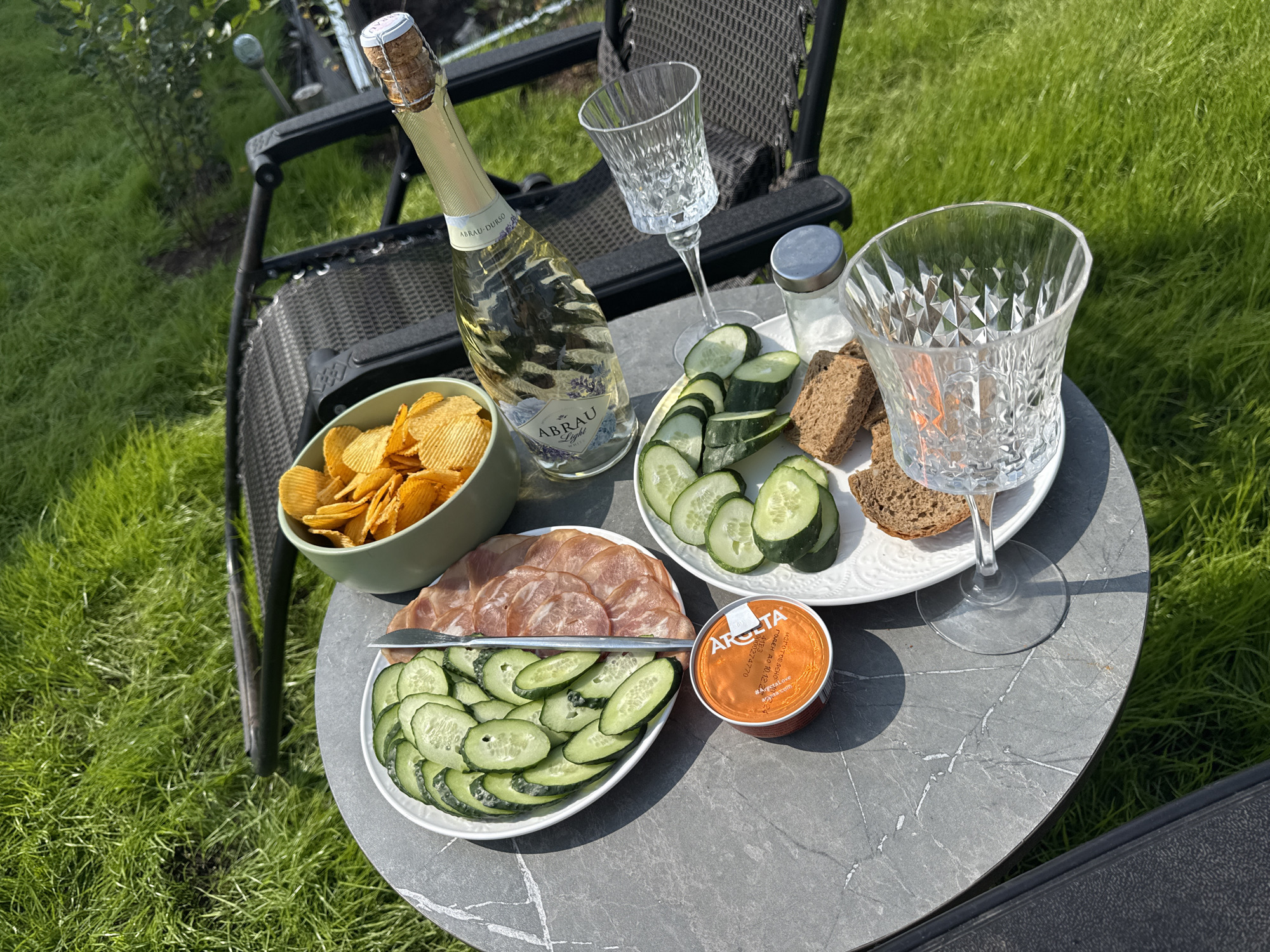 An outdoor picnic is set up on a table in a grassy area. A bottle of champagne, two crystal glasses, a bowl of potato chips, sliced cucumbers, assorted cured meats, and pieces of bread are arranged on a marble-patterned round table. The scene is set in Лобаново, Russia, with lush green grass and foliage visible in the background. The lighting suggests it is daytime, with sunlight casting shadows. A portion of a black outdoor chair is also visible.