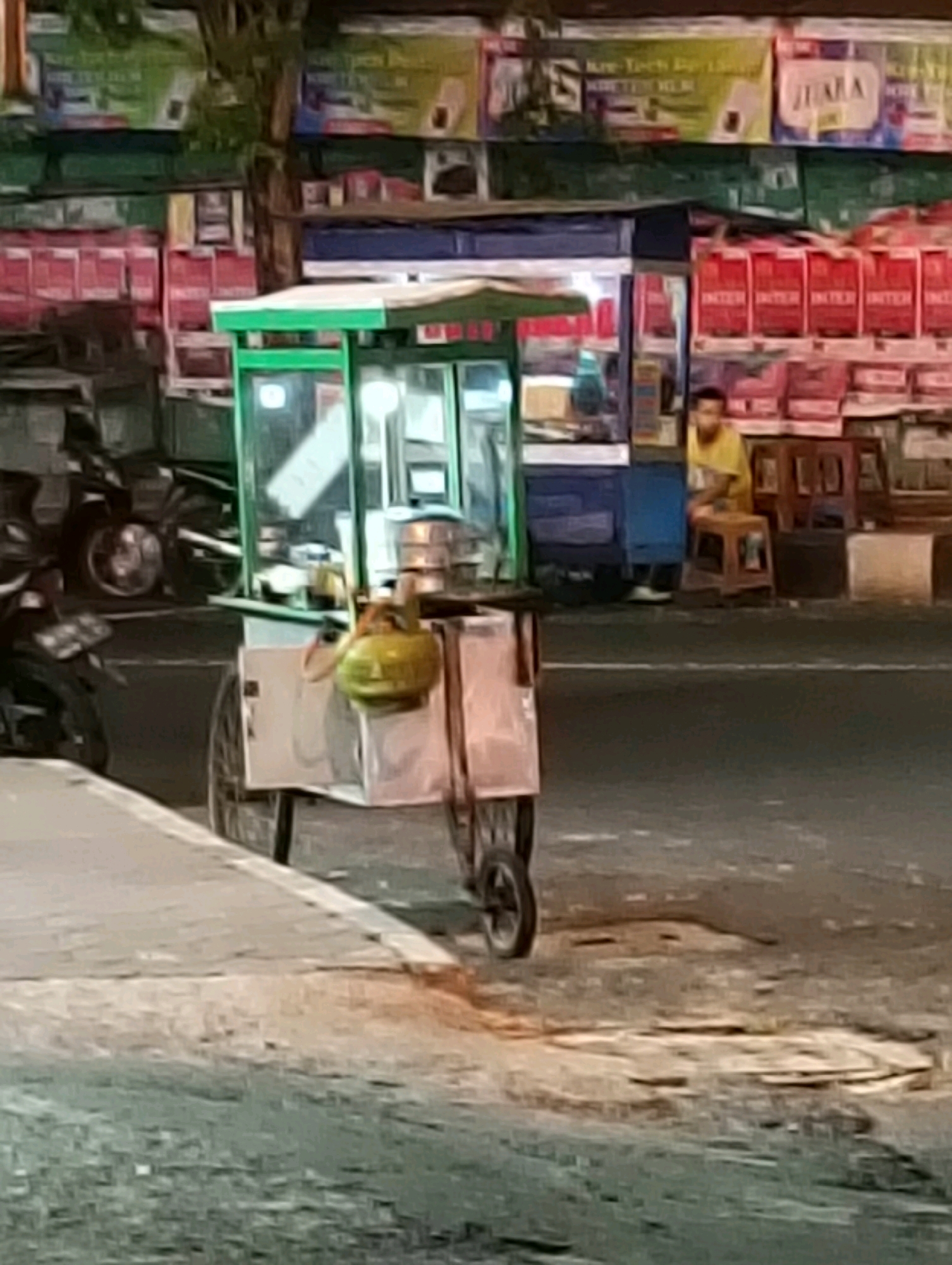 The image depicts a street scene at night in Purworejo, Indonesia, near a food stall. A street food cart with a green frame and glass enclosure is prominently featured in the foreground, equipped with a yellow gas canister. To the left, a motorcycle is partially visible, suggesting it is parked or passing by. In the background, a man wearing a yellow shirt sits on a stool outside a blue stall, with shelves of products behind him. The environment includes a paved sidewalk, a street, and several commercial stalls with various goods displayed. The lighting indicates it is nighttime, with artificial lights illuminating the area.