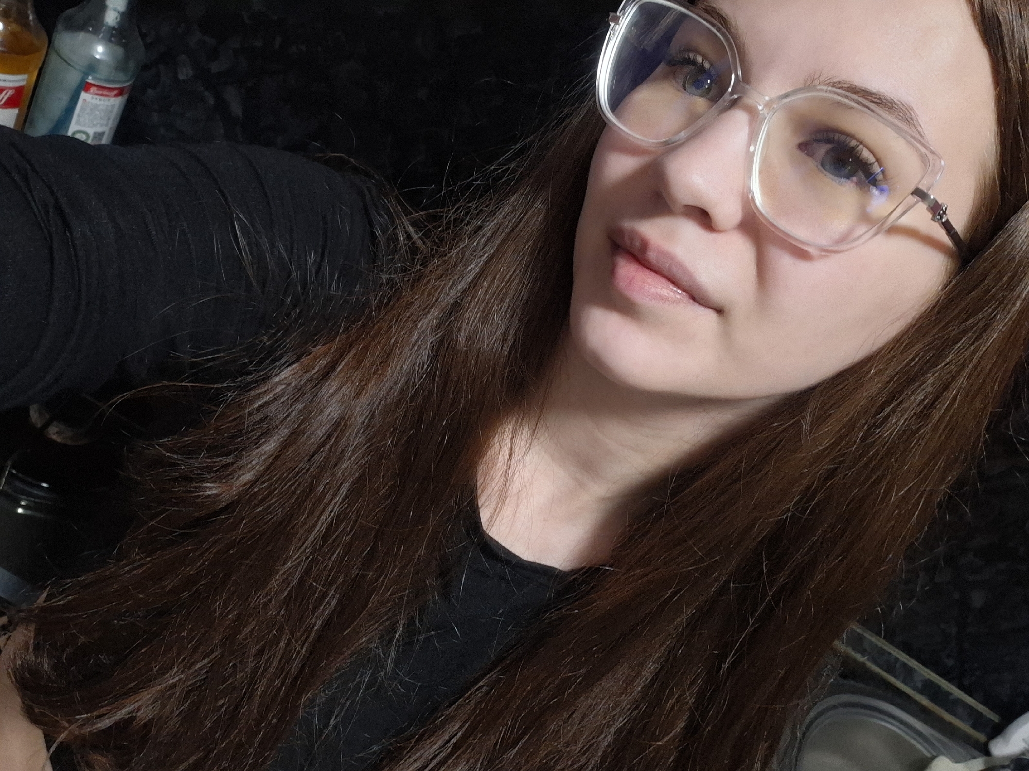 A selfie shot shows a young woman with long, brown hair wearing clear-framed eyeglasses. She is wearing a black shirt. Bottles are visible in the upper left corner of the frame. The location is possibly Chaikovsky, Russia, although this cannot be confirmed from the image.