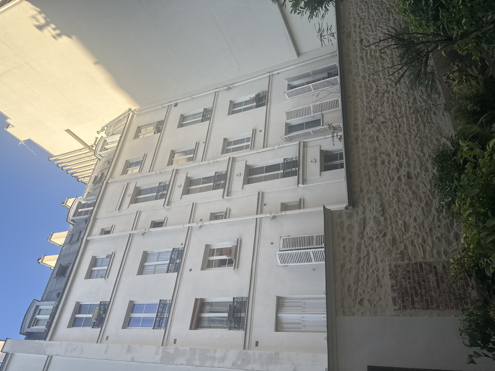 This image depicts a narrow courtyard in Paris, France, with an old, pale yellow apartment building dominating the frame. The building exhibits classic Parisian architecture, featuring multiple stories with numerous windows. Many of the windows have white shutters, some of which are open, revealing interior spaces. The lower part of the courtyard is enclosed by a rough stone wall, with a section of exposed red brick visible. Green foliage and plants are present at the bottom of the frame, suggesting a garden area. Sunlight casts a shadow from an adjacent, larger building onto the right side of the frame, indicating a daytime scene. The sky above is a clear, bright blue.