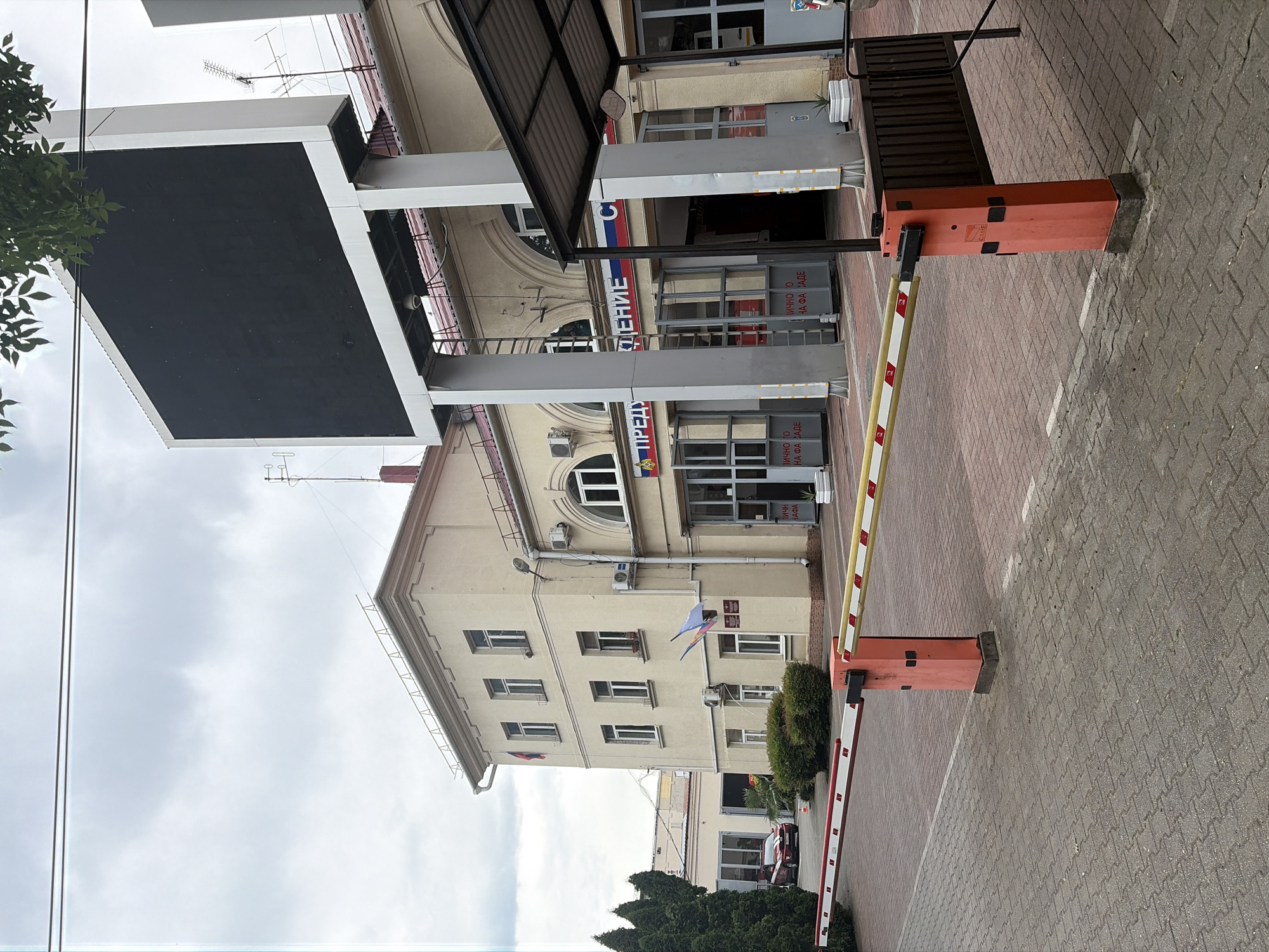 The image depicts the entrance to a building in Sochi, Russia, as evidenced by the Cyrillic signage which reads "ПРЕДУПРЕЖДЕНИЕ" (warning) and "СП" (likely an abbreviation for "Спасательная часть" or rescue unit). The building appears to be a public service facility, possibly a fire station, given the presence of emergency vehicle bays visible through the large glass doors. A red car, possibly an emergency vehicle, is parked to the left of the building. A retractable barrier gate, painted with red and yellow stripes, is positioned across the paved entrance, suggesting controlled access to the premises. The sky is overcast, indicating a cloudy day. The foreground consists of a paved courtyard with a brick-patterned surface. The overall scene conveys a sense of official premises with security measures in place.