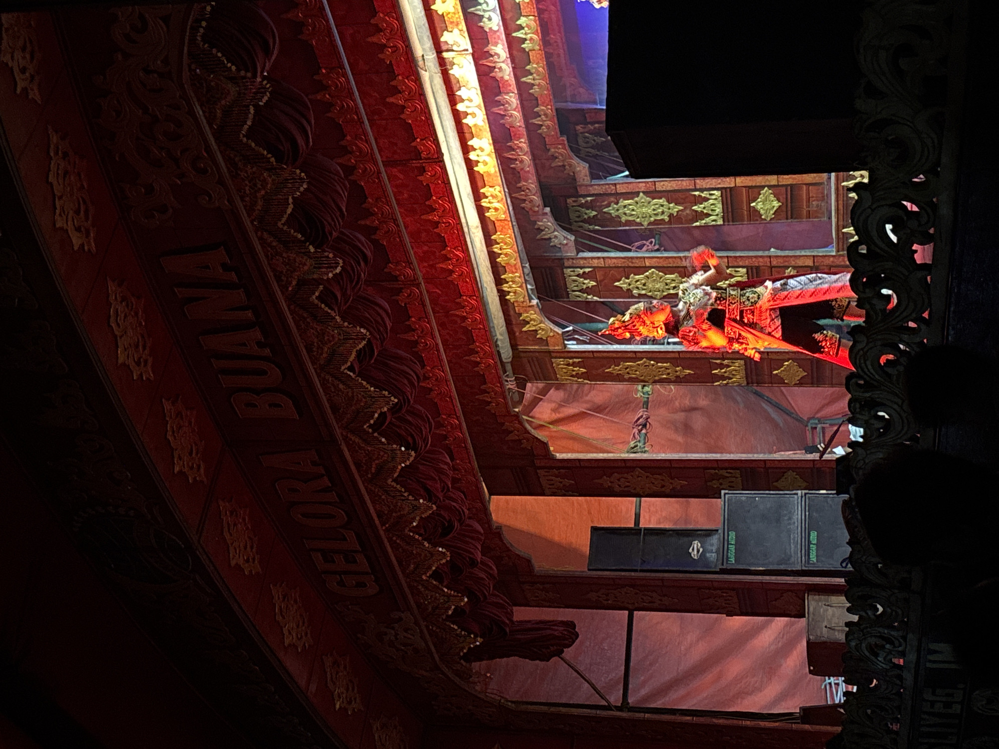 The image shows a performance on a stage in Panongan, Indonesia. Above the stage, a large sign reads "GELORA BUANA" in stylized, golden lettering against a red background. The stage itself is decorated with ornate red curtains and golden trim. In the center of the stage, a performer in a vibrant, traditional costume, including a headdress and flowing red cape, is illuminated by red stage lights. The performer appears to be in motion, possibly dancing or acting. To the left of the stage, two large black speaker cabinets are stacked on top of each other, each with a label that reads "LANGGAN AUDIO." A wooden crate and part of a white fabric backdrop are also visible on the left side of the stage. The foreground shows the edge of a seating area, with darkened silhouetted figures of audience members and decorative wooden railings. The overall atmosphere is dramatic and theatrical, with the red lighting and elaborate decorations creating a sense of spectacle.