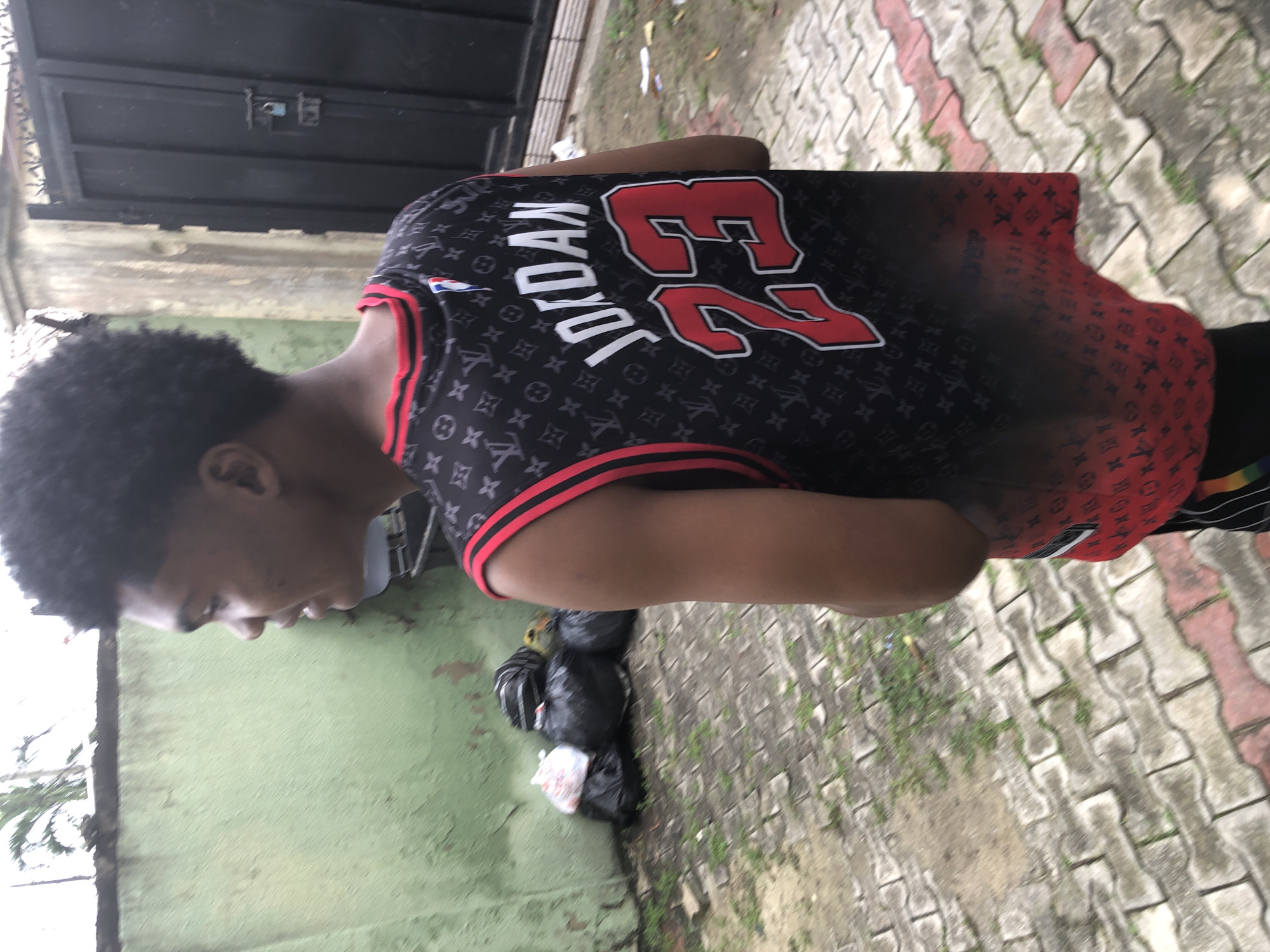 A young male is walking away from the camera, wearing a black and red basketball jersey that says "JORDAN 23" on the back. The jersey has a repeating Louis Vuitton monogram pattern. He is in an outdoor setting with a light-colored, paved ground made of interlocking bricks. To his left, there is a weathered, green painted wall with some graffiti or peeling paint. In front of the wall are several black trash bags and one white trash bag. Behind him, to the right, is a tall black metal gate with a padlock. The environment appears to be an urban setting in Ado, Nigeria, possibly a residential or commercial area, given the mixture of residential elements like the gate and wall with the presence of trash. The sky is overcast.