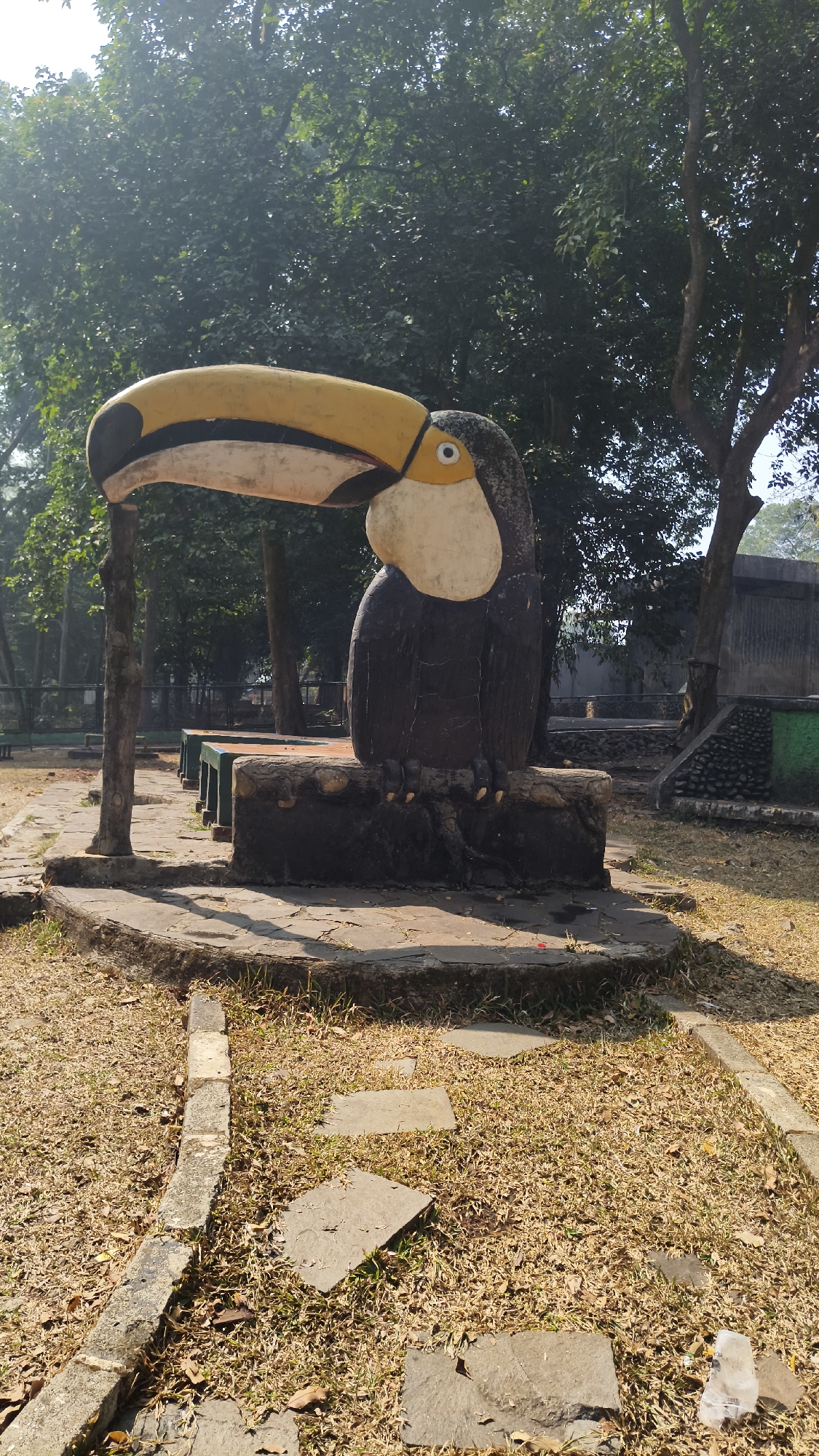 A statue of a toucan is situated in an outdoor park area in South Jakarta, Indonesia. The statue is dark brown with a large, curved yellow beak accented with black. The toucan is perched on a rough, log-like base, which rests on a circular stone platform. In the background, several trees with green foliage are visible. To the left of the toucan statue, a row of empty green benches with brown seats is arranged. The ground is covered with dry, brown grass and scattered flat stones, forming a pathway. A low stone border runs along the edge of the pathway. The overall environment suggests a public park or zoo enclosure.