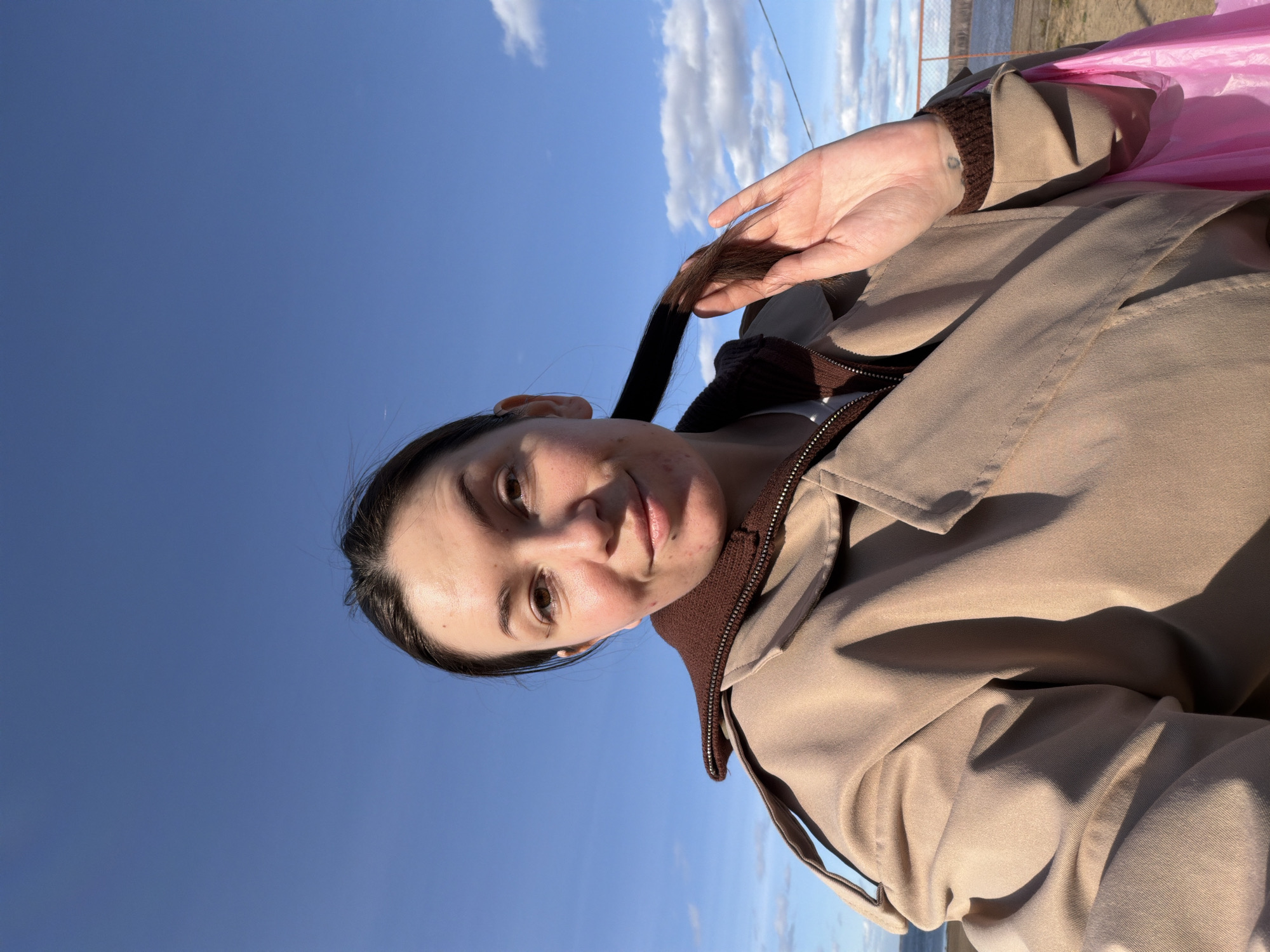 A selfie taken outside in Chaikovsky, Russia, shows a woman with dark hair pulled back in a ponytail, wearing a tan jacket with brown trim, against a background of blue sky and clouds. She is holding the end of her ponytail in her hand. A body of water is visible in the background. A pink object is next to her.