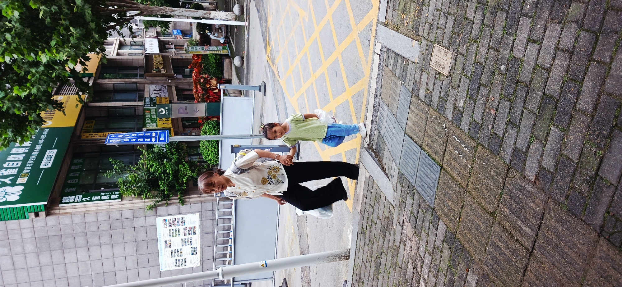 An elderly woman and a young girl are holding hands and walking on a sidewalk in Jiulongpo District, China. The sidewalk is made of dark gray brick and abuts a street painted with yellow crosshatch lines. The woman wears a light-colored shirt with floral designs and black pants. The girl wears a light green shirt and blue pants. Buildings and trees are visible in the background, along with signs and street furniture.