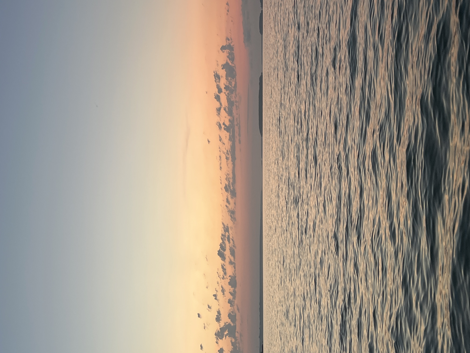 A serene view of a body of water under a gradient sky at sunset. The sky transitions from a pale blue at the top to soft oranges and pinks closer to the horizon, dotted with small, fluffy clouds. The water, appearing calm with gentle ripples, reflects the warm colors of the sky. In the distance, along the horizon, are faint outlines of landmasses, possibly islands or a coastline. The overall scene suggests a peaceful, natural setting. The image does not contain any people, specific objects, or actions that would indicate a city location.