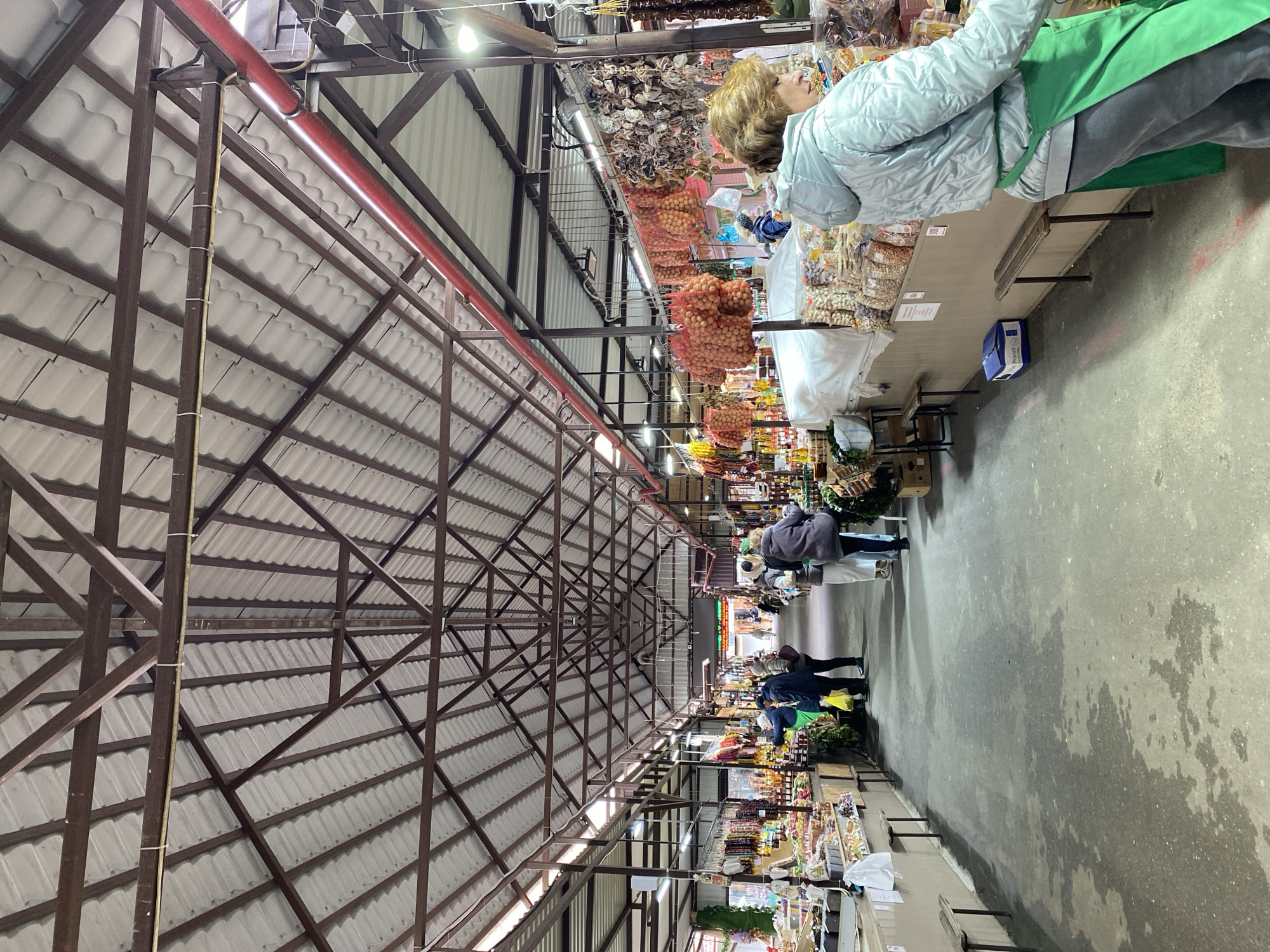 The image shows the interior of a covered market in Gelendzhik, Russia. The market is long and narrow with a high, peaked roof supported by a brown metal framework. The roof is made of overlapping white tiles. A thick red pipe runs along the length of the ceiling.
Market stalls line both sides of a wide walkway. The stalls are filled with various goods, including dried fruits, nuts, and other food items. People are browsing and shopping at the stalls. In the foreground, a woman with blonde hair is wearing a light blue jacket and a green apron while standing behind a stall. Further down the walkway, several shoppers are walking. The floor is a gray concrete surface. The lighting is somewhat dim, with overhead lights providing illumination.