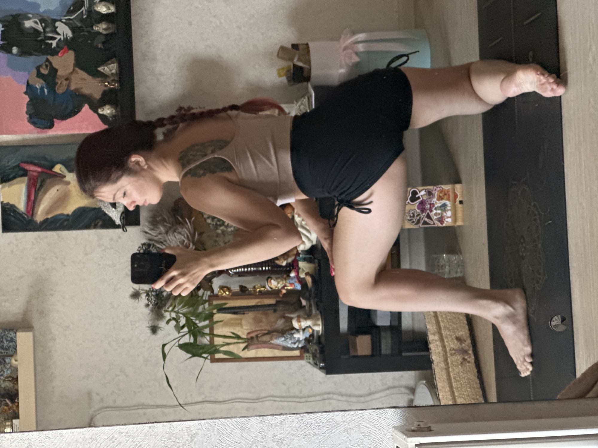 A young woman with braided dark red hair is captured in a mirror reflection, performing a low lunge yoga pose. She is wearing a light beige sports bra and black drawstring shorts. The background reveals a room with a textured wallpaper, a black shelf filled with decorative items including plants, figurines, and artwork featuring comic book characters. A yoga mat is visible on the floor. The scene is set in an indoor environment, likely a home.