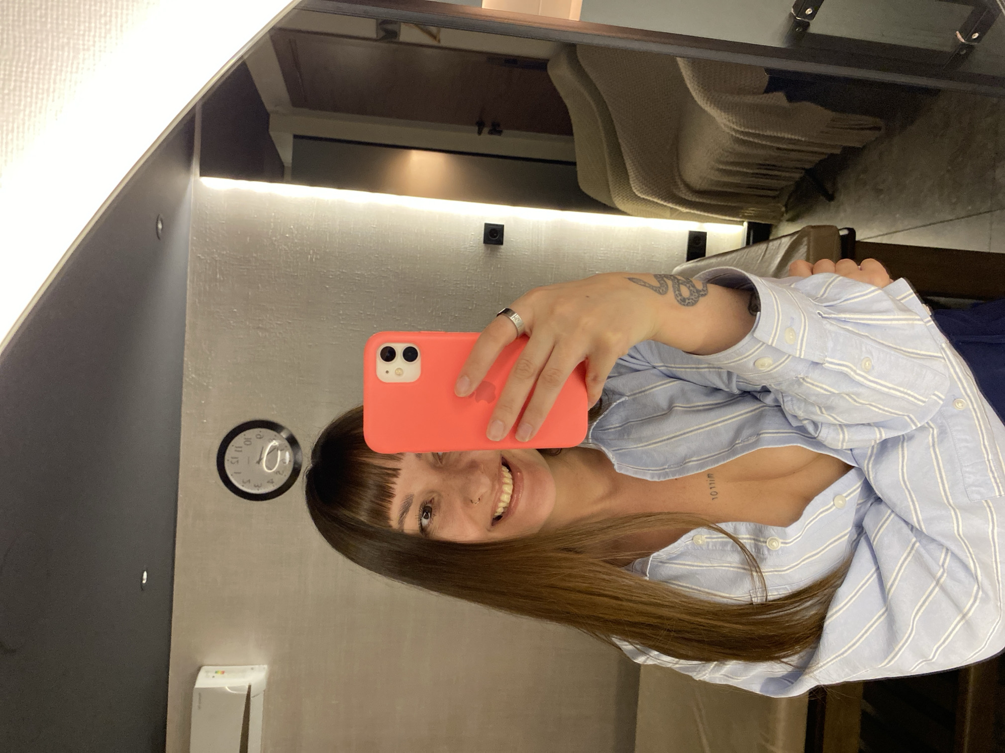 A woman is taking a selfie in a mirror. She is holding a coral-colored iPhone in front of her face. She has brown hair with bangs and is wearing a blue and white striped shirt. A clock on the wall next to her displays the time. The location may be in the city of Votkinsk, Russia.