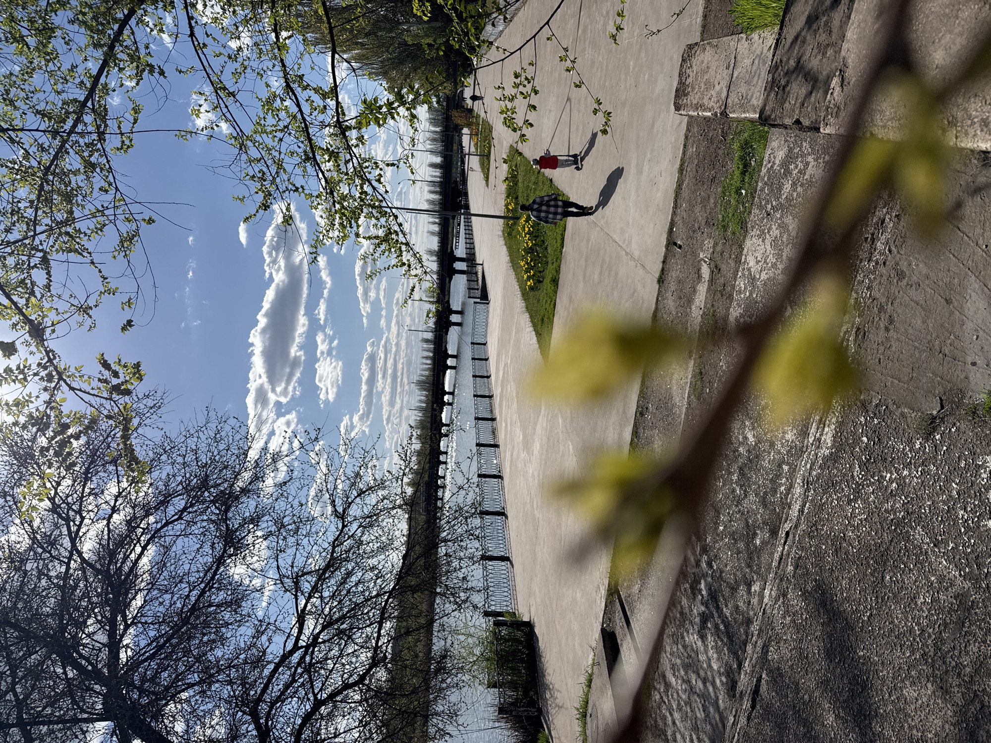The image presents a view of a waterfront promenade in Kryvyi Rih, Ukraine, under a partly cloudy sky. A concrete pathway runs parallel to a body of water, with a decorative metal railing separating the two. Two individuals are present on the path: one wearing a red shirt riding a scooter, and another dressed in a plaid shirt walking. The promenade is bordered by grassy areas with patches of yellow flowers. In the foreground, tree branches with fresh green leaves are partially obscuring the view. The sky is blue with elongated white clouds. On the right side of the image, there is a concrete barrier with shadow patterns. Bare trees are on the left.