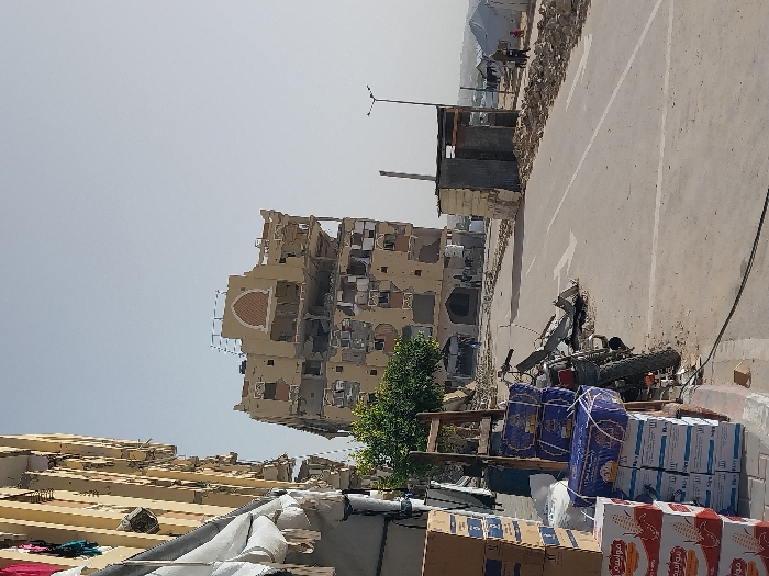 This image depicts a scene of extensive urban damage in Khan Yunis, Palestinian Territory, under a clear, light blue sky.
In the foreground, on the left, a tall, light-colored building, possibly residential, shows visible exterior damage and has laundry hanging from some balconies. On the sidewalk below, stacks of cardboard boxes, some labeled "مواسيه" (Mowasah), are present, alongside a partially damaged motorcycle lying on its side. A wooden A-frame structure leans against the building. The asphalt road in the mid-ground has a white dividing line and a white directional arrow painted on it, with debris, rubble, and dirt scattered along its edges.
Dominating the center-background is a multi-story building, light tan in color, which has sustained severe structural damage. Multiple floors show exposed interiors, shattered windows, and large sections of the facade missing, revealing concrete and rebar. The top floor retains an arched decorative element. A small, verdant tree stands in front of this damaged building. Two indistinct figures are visible near its base.
To the right of the road, a small, makeshift structure made of corrugated metal and concrete blocks stands. Beyond this, the terrain becomes unpaved and dusty, leading to a visible displacement camp comprised of several white tents. Three figures—two adults and one child—are walking on the right side of the road in the direction of the tent camp. A solitary streetlamp stands further down the road on the right.