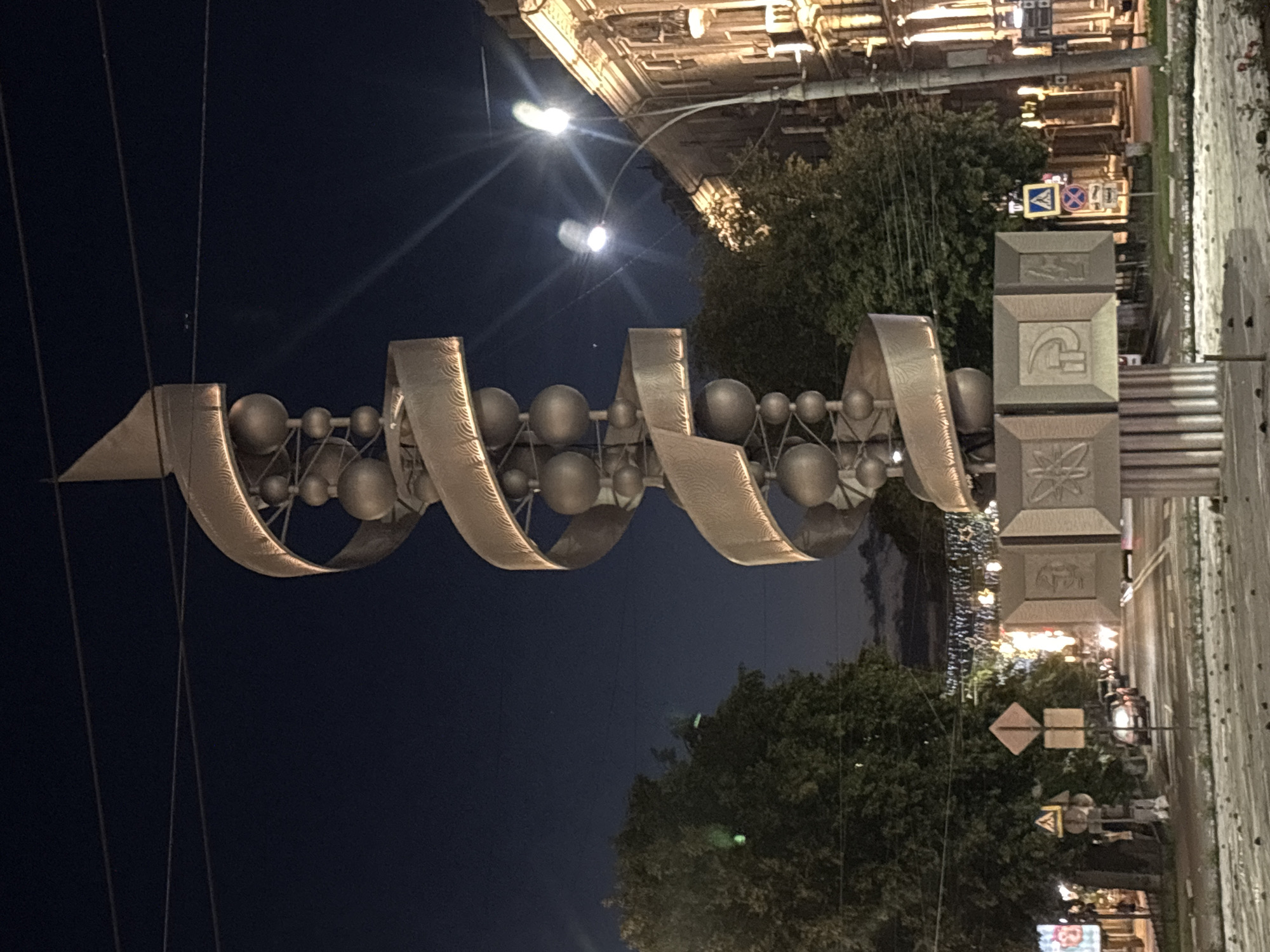 The image shows a nighttime view of a modern sculpture in Novaya Usman', Russia. The sculpture is tall and spiral-shaped, with metallic bands and spherical elements. At the base of the sculpture are square panels with embossed symbols. The sculpture is illuminated, casting a glow on its surroundings.
The environment is urban. Trees line the street, and buildings with illuminated windows are visible in the background. Streetlights and traffic lights are present, indicating a street scene. Several cars are on the road, and some traffic signs are visible. The sky is dark, suggesting it is nighttime. The ground in the foreground appears to be a paved surface, possibly a street or plaza, with some debris scattered on it. There are no people clearly visible in the image.