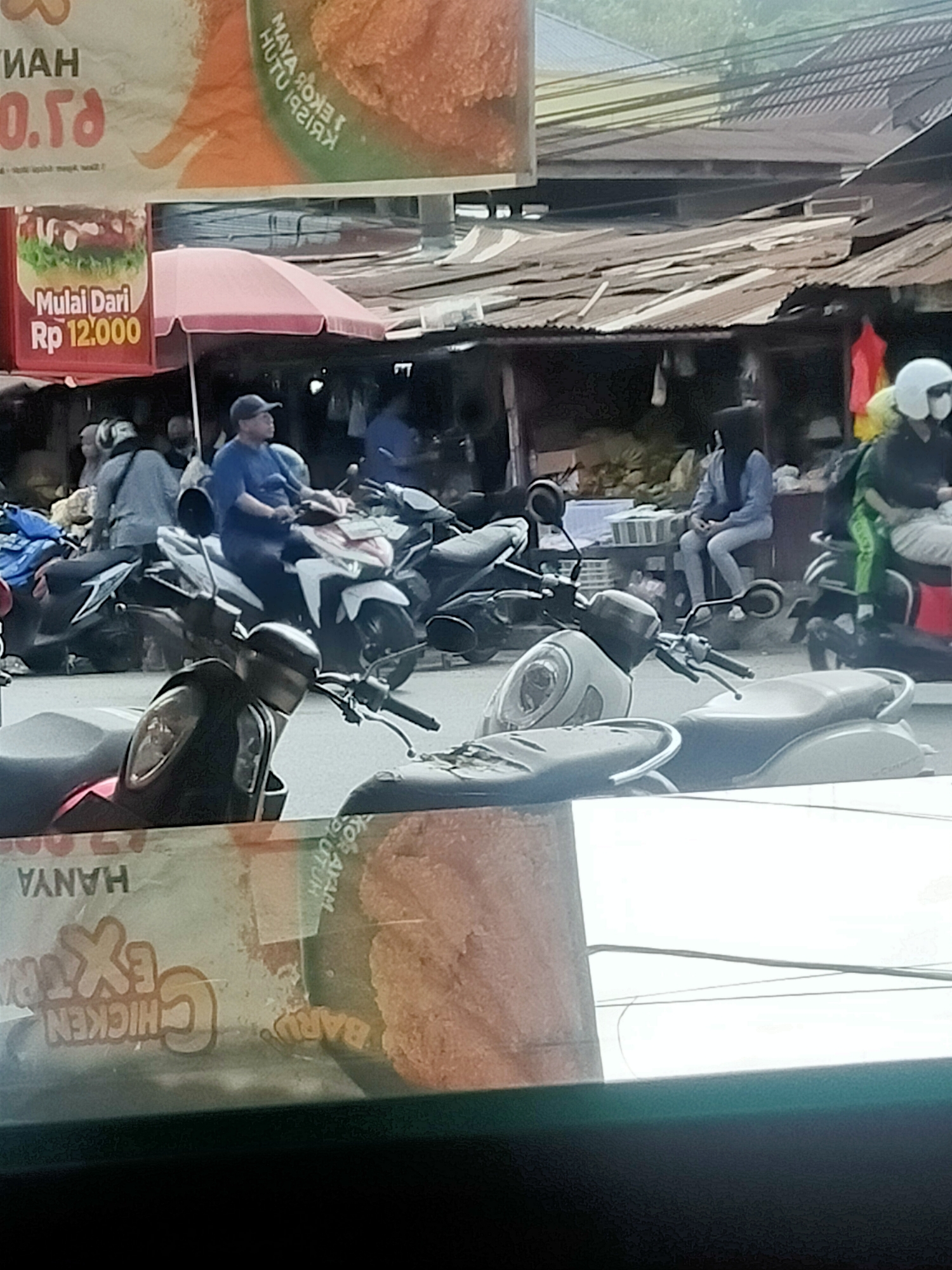 The image shows an outdoor scene with several people on motorcycles in a market or street setting. In the foreground, there are two parked motorcycles, one black and red, and another light-colored scooter. Behind them, a man in a blue shirt and a black cap is seated on a white motorcycle. Several other motorcycles are visible further back, along with people standing or sitting around them. The background includes makeshift stalls with corrugated metal roofs, some of which are selling goods. There are also banners with text and images, including one that appears to be an advertisement for chicken. The overall environment suggests a busy, informal commercial area.