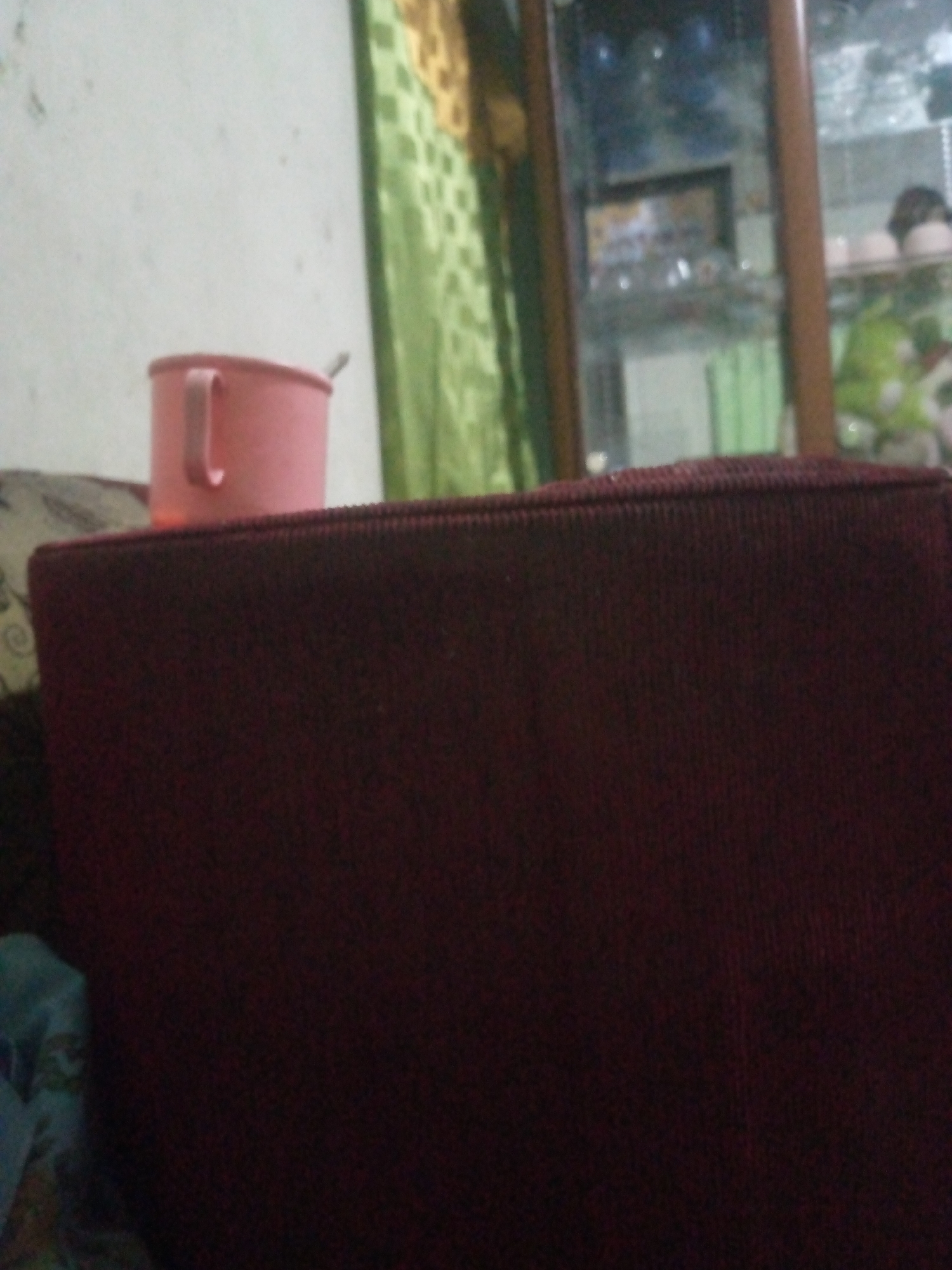 A pink mug with a handle is visible on the left side of the image, placed on what appears to be a dark red, textured surface. A spoon is sticking out of the mug. Behind the mug, a green patterned fabric is partially visible. To the right, a glass-fronted cabinet is seen, reflecting light and displaying items within, including what appear to be toys and other objects. The overall scene suggests an indoor setting, possibly a domestic environment in Laksanamekar, Indonesia. There is no explicit indication of any activity or interaction taking place.