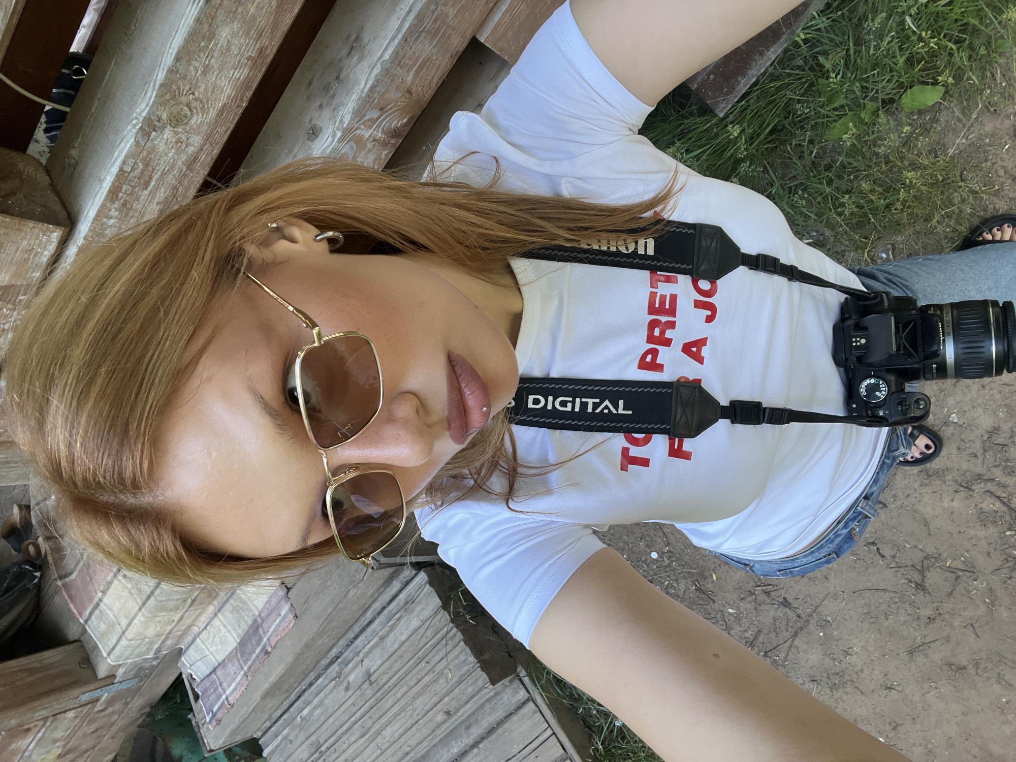 A young woman with light brown hair, wearing sunglasses, a white t-shirt with red text, blue jeans, and sandals, is taking a selfie outdoors. She has a lip piercing and is wearing hoop earrings. A camera hangs around her neck, and she is standing on a dirt and grass area next to wooden structures. The environment suggests a casual outdoor setting, possibly a park or a backyard. The presence of wooden planks and grass indicates a natural or semi-natural environment. No specific landmarks or elements definitively place this image in Chaikovsky, Russia, other than the stated location.