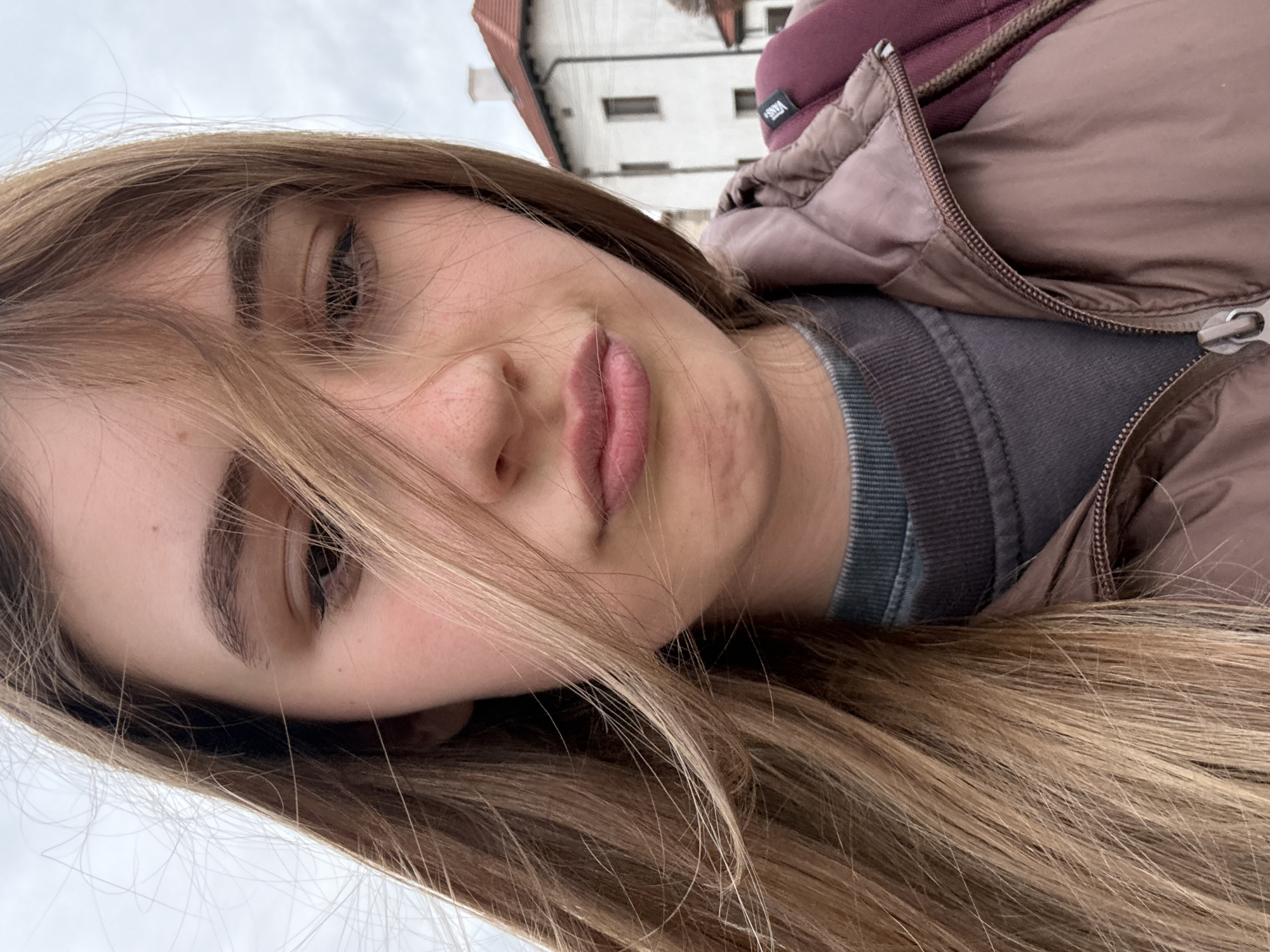 A close-up selfie captures a young woman outdoors, rotated to appear sideways. Her light brown hair cascades across her face, partially obscuring it, and her visible eye looks towards the camera. She has light skin, dark eyebrows, and full pink lips held in a slight pout. She is dressed in a dark gray ribbed sweater layered under a light brown puffy jacket. A burgundy fabric, likely a hood or backpack strap, is visible near her shoulder, featuring a white tag with the brand name "VANS". The background is softly blurred, showing a white building with a reddish-brown tiled roof against a bright, overcast sky. The scene is set in Panagyurishte, Bulgaria, on what appears to be a cool, cloudy day.