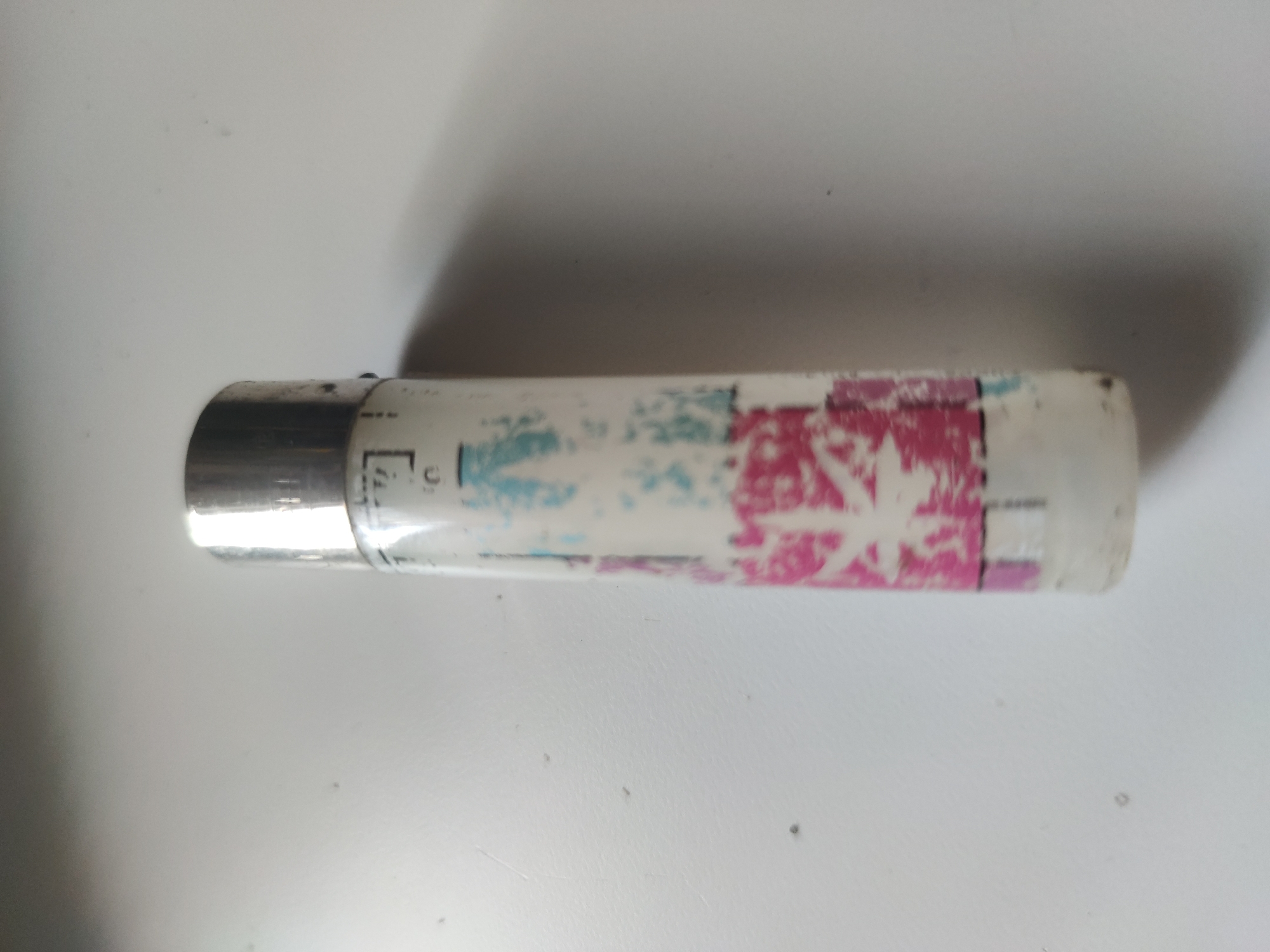 An object is lying horizontally on a white surface. The object is cylindrical, with a metallic end and a white body featuring faded blue and pink markings. The location is Unknown, Unknown.