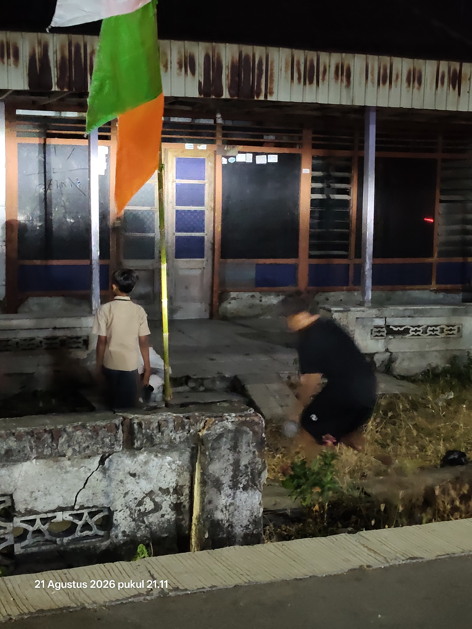 In the city of Wedi, Indonesia, an image captures a scene at night. A young person, seen from behind, stands facing a weathered building. They are wearing a light-colored shirt and dark pants. To the right, another figure, largely obscured by motion blur, appears to be engaged in an activity near overgrown vegetation. In the foreground, a flag with green and orange sections hangs from a pole, and a cracked concrete structure is visible. The timestamp at the bottom of the image reads "21 Agustus 2026 pukul 21.11".