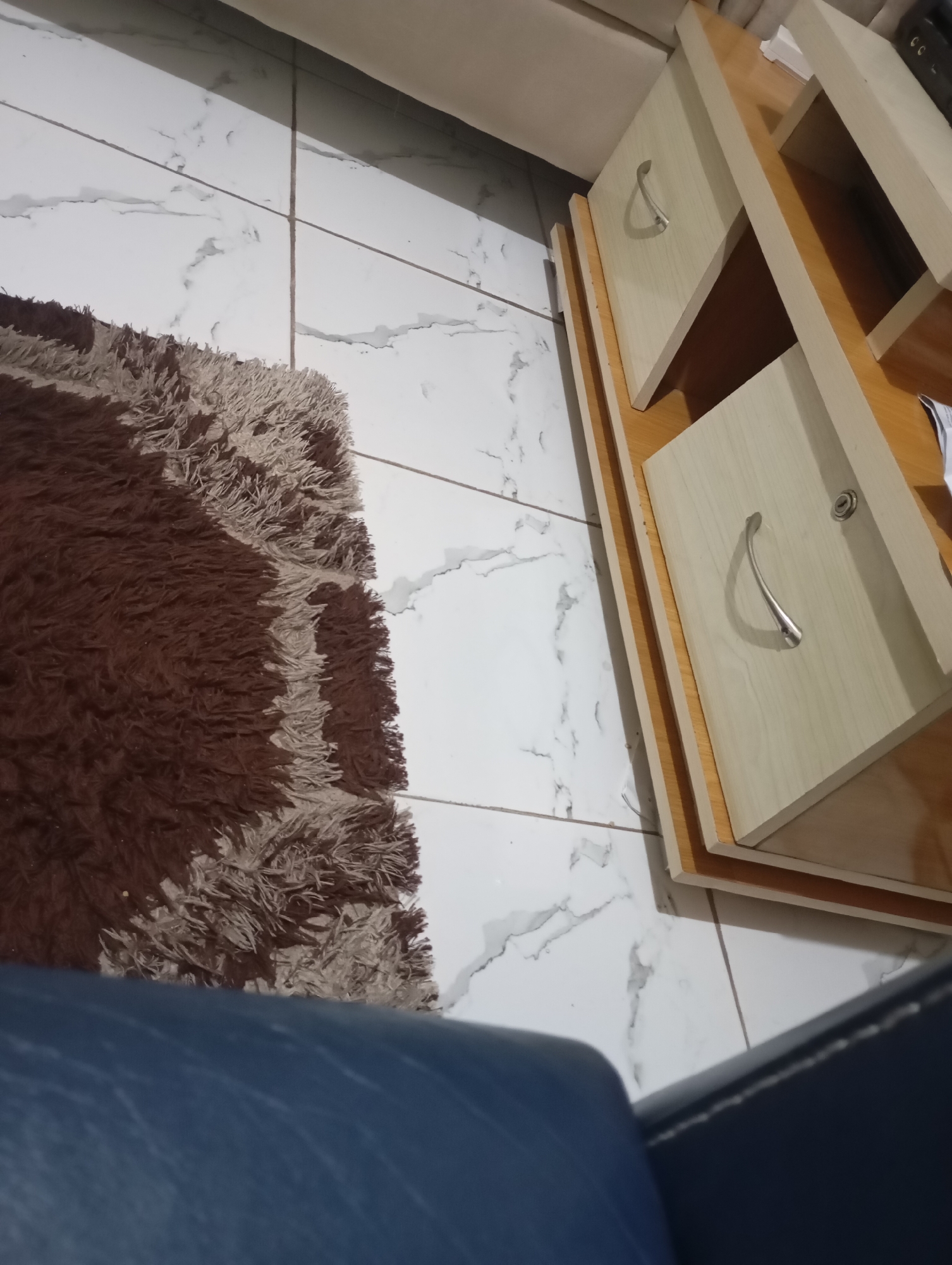An overhead, slightly angled view shows a fluffy, brown and beige rug on a white tiled floor. To the right, a light-colored wooden cabinet with two open drawers is visible. Each drawer has a silver handle. The cabinet is open and empty. The scene is dimly lit, casting shadows from the cabinet onto the floor and rug. A dark blue upholstered object, possibly a couch or chair, is in the lower portion of the image, partially obscuring the rug and floor. The image is devoid of any people or obvious signs of activity. The location is identified as Unknown, Unknown.