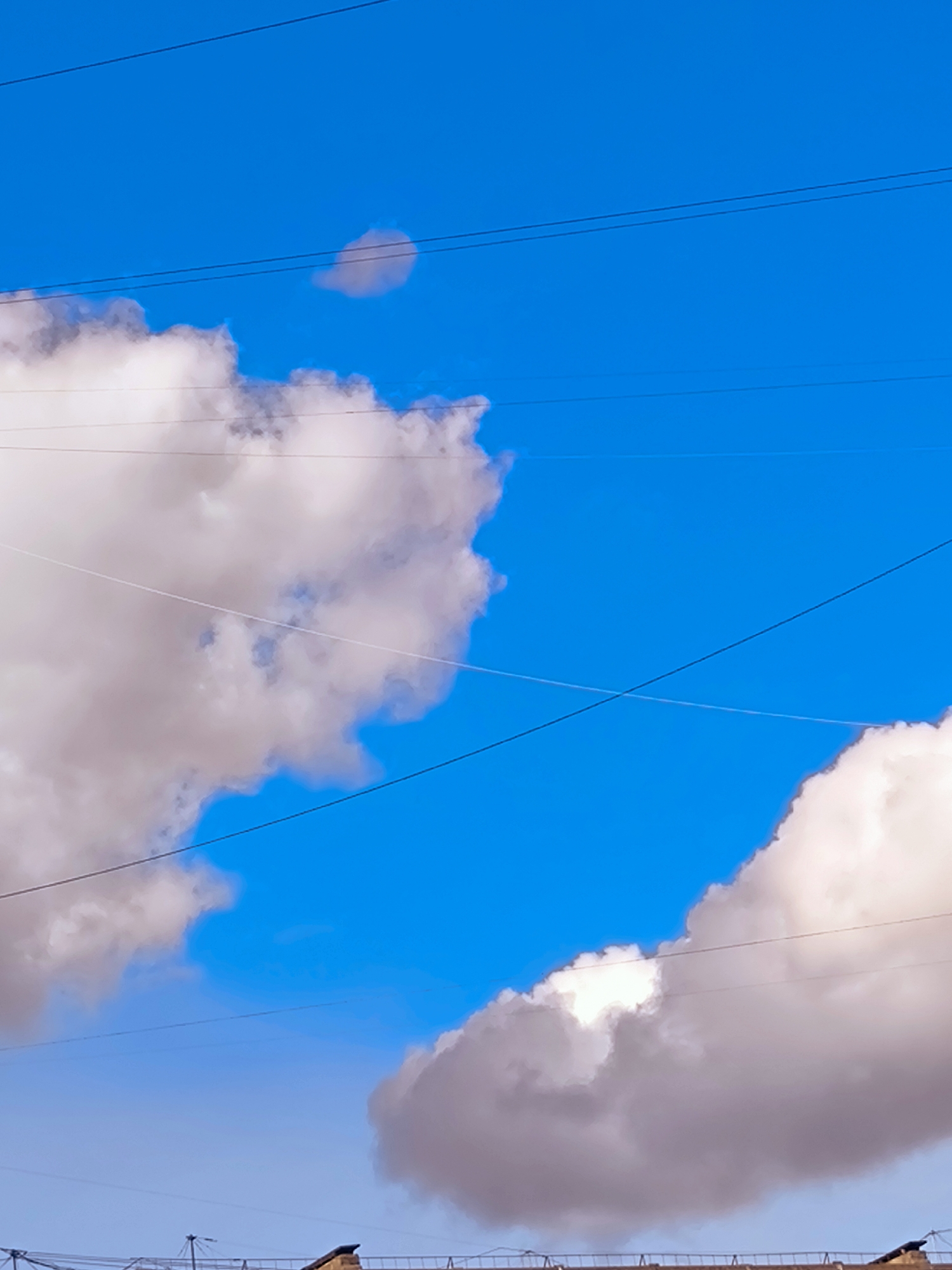 The image depicts a bright blue sky with scattered clouds, crisscrossed by several power lines. The clouds are predominantly white and light pink, suggesting they are illuminated by sunlight. There's a faint, small cloud visible in the upper center of the frame. In the lower portion of the image, the rooftops of buildings can be seen, hinting at an urban setting. This scene is consistent with a view from a high vantage point in the city of Riga, Latvia, possibly a balcony or rooftop overlooking the city.