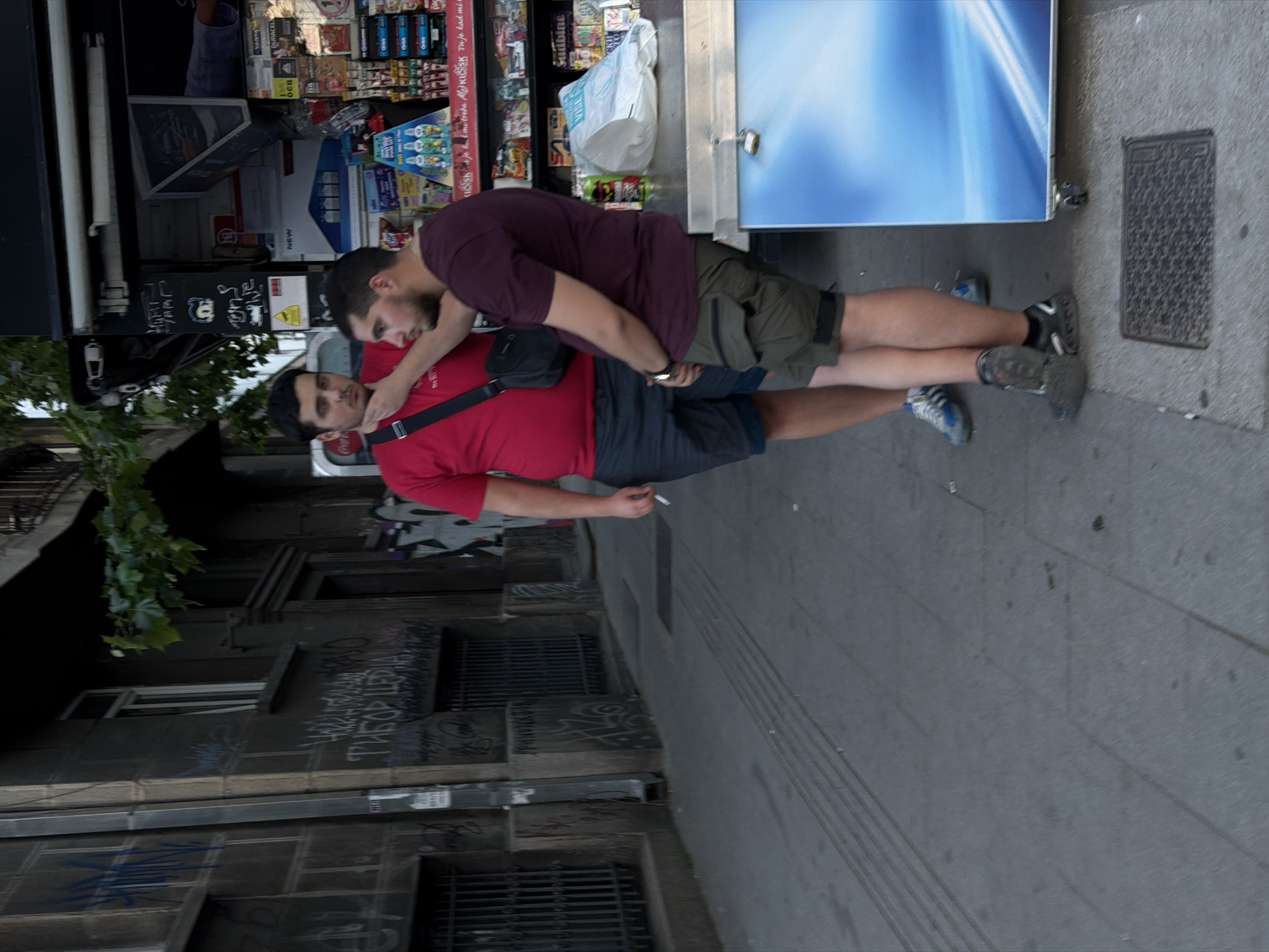 Two men are standing outside a kiosk on a street in Belgrade, Serbia. One man is wearing a red shirt and the other is wearing a purple shirt. The man in the purple shirt has his arm around the other man's neck. The man in the red shirt is holding a cigarette. The kiosk is stocked with various items. The building adjacent to the street has graffiti on the wall.