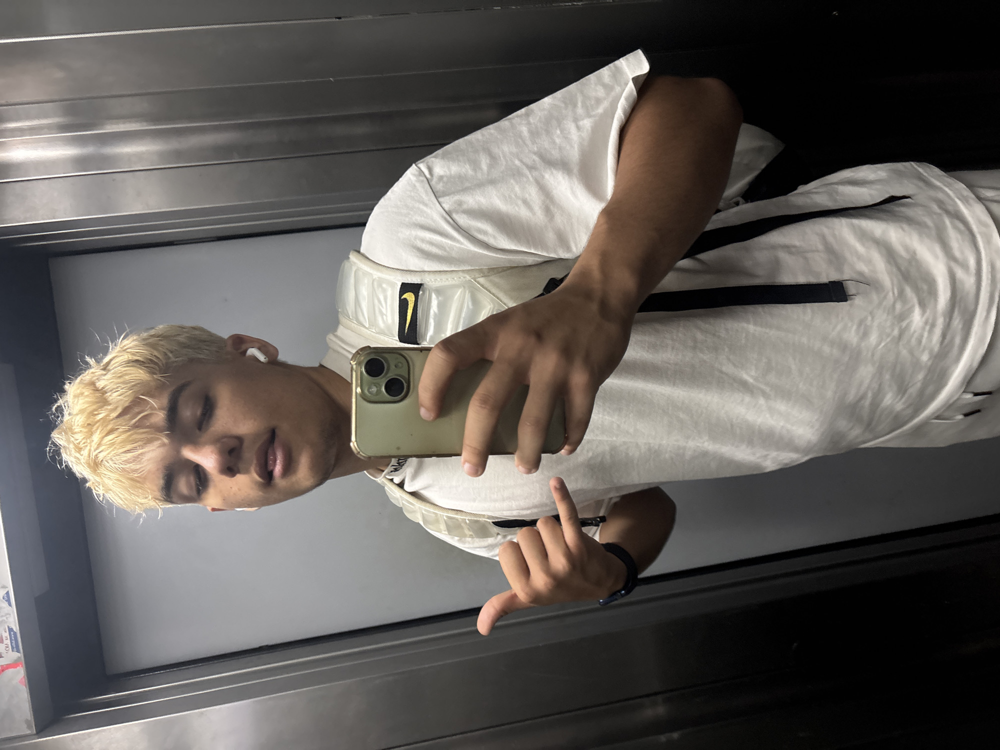 A young person with bleached blonde hair is taking a selfie in an elevator. They are wearing a white t-shirt and a white backpack with a black strap. The person is holding a smartphone in their right hand and making a hand gesture with their left hand. The elevator walls are made of brushed metal, and there is a grey door in the background. The person is also wearing white earbuds. The lighting is overhead and somewhat dim. There are some stickers on the ceiling of the elevator. It is not possible to determine the location within Sofia, Bulgaria from the image.