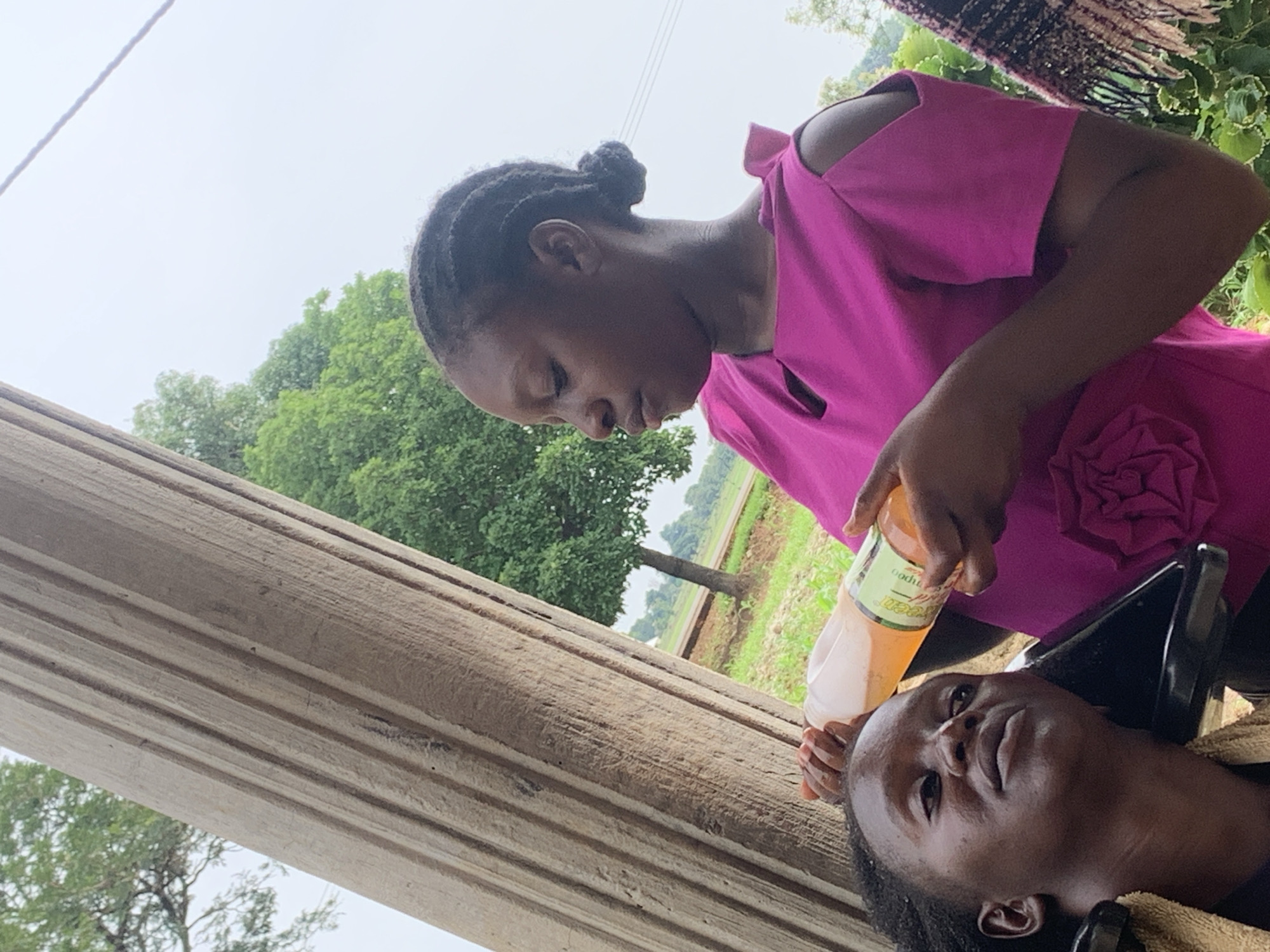 In Agban, Nigeria, a young woman with dark skin and braided hair styled in a bun, wearing a bright pink off-the-shoulder top adorned with a fabric flower, is holding a clear plastic bottle of amber-colored liquid. She is pouring the liquid onto the head of another young woman who is reclined in a black salon chair, wearing a tan towel around her neck. The background features a large, textured, wooden post on the left and lush green trees and an overcast sky on the right.