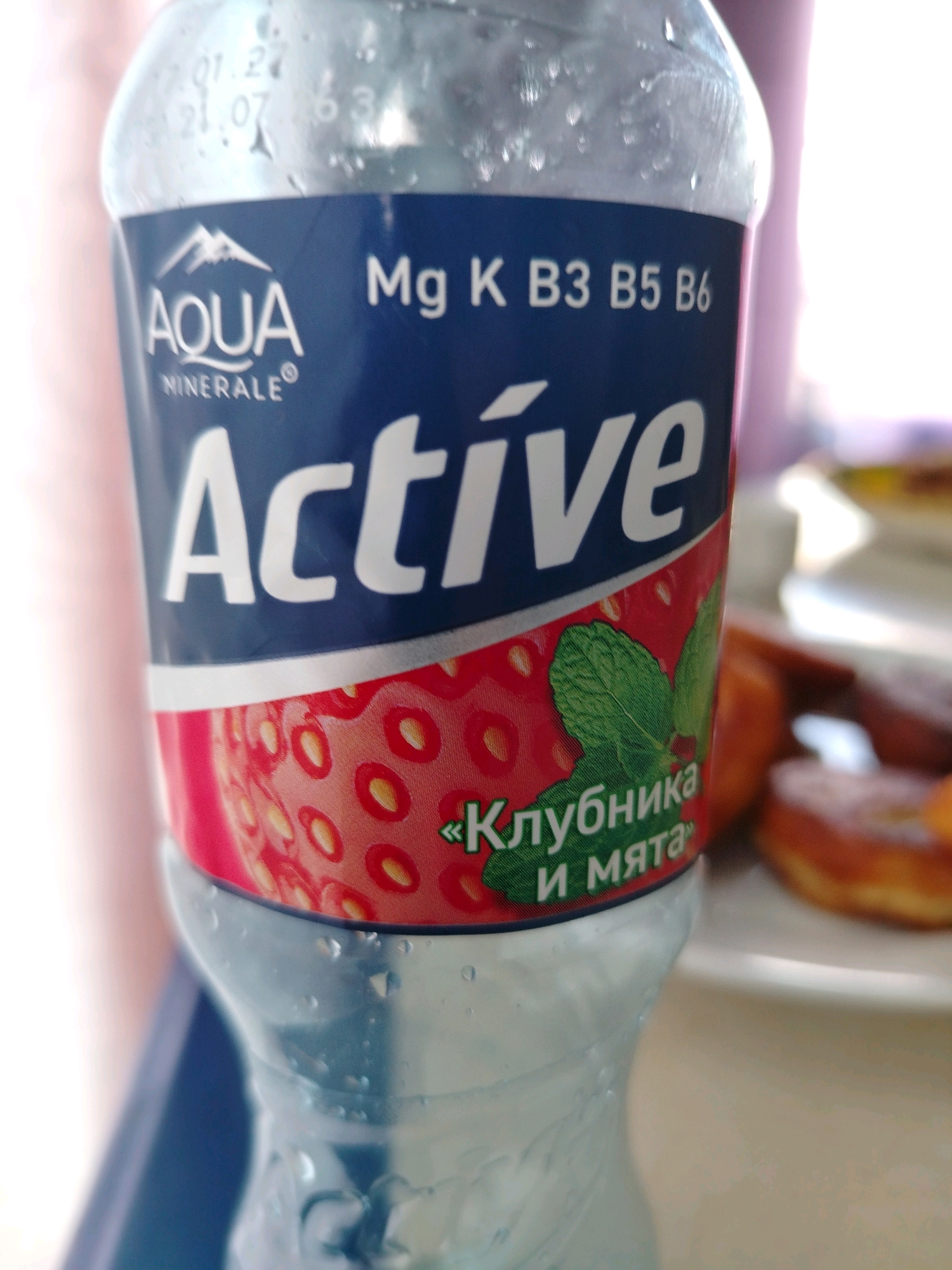 A close-up, slightly out-of-focus shot shows the label of a plastic water bottle. The label is primarily dark blue with white text. Prominently featured is the word "Active" in large white letters. Above "Active," the word "AQUA" is visible in white text with a mountain logo. To the right of "AQUA," the text "Mg K B3 B5 B6" is also in white. The lower part of the label displays a red design with white speckles, resembling strawberries, and a green mint leaf. Text in Russian, including "Клубника" and "и мята," is visible on this lower section. The bottle itself is clear plastic with condensation droplets on its surface. In the background, out of focus, are soft pink and white areas, and to the right, blurred shapes of food items that appear to be donuts or pastries. There is no discernible information about the city of Pregradnoye, Russia in the image.