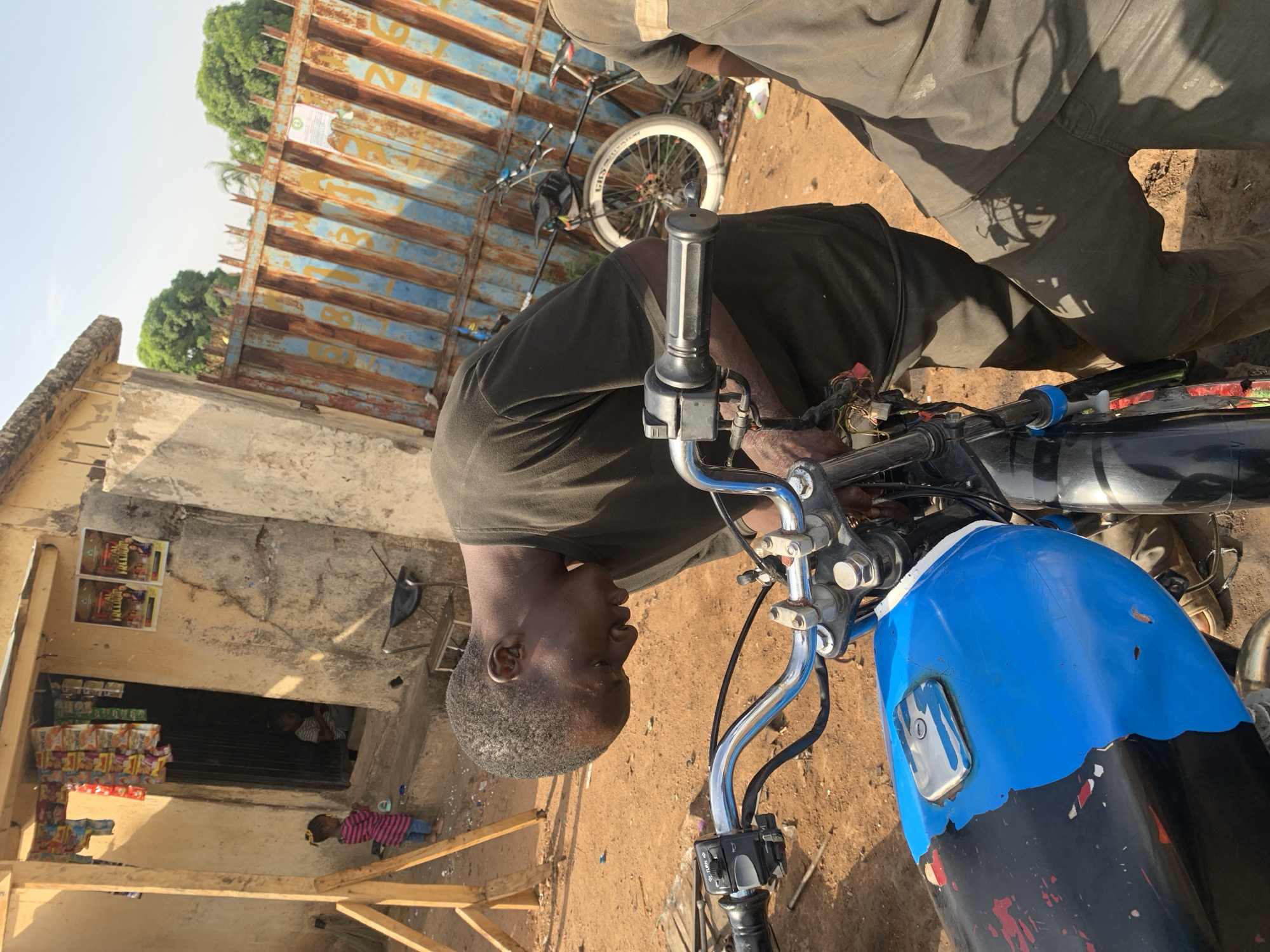 A man is working on a blue motorcycle in Gura Top, Nigeria. He is bent over the handlebars, facing left. Another person is standing nearby, with their legs and lower body visible in the upper right. The scene is set outdoors on a dirt surface. In the background is a building with a storefront and goods on display, with a child standing in front. A rusty metal structure is visible behind the building.