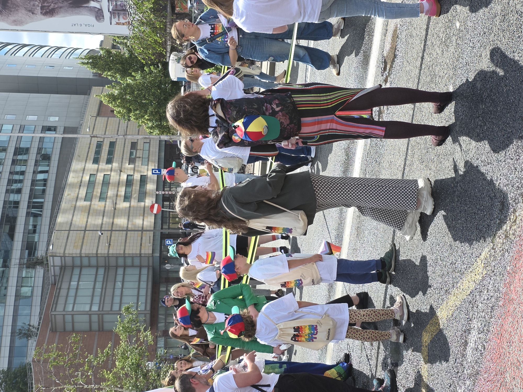 The image captures a daytime outdoor scene in Warsaw, Poland, likely during a Pride event, as evidenced by the presence of rainbow flags and attire. A group of people are gathered on a street, with modern buildings forming the backdrop.
In the foreground, several individuals are visible. Two young children, one on the left and one in the center, are wearing white t-shirts and light-colored backpacks adorned with rainbow designs and the "Deloitte" logo. Both children are wearing matching baseball caps with colorful panels. To the right of the children, a person with curly brown hair wears a multi-colored striped dress and a patterned backpack with a colorful baseball cap attached. Further to the right, a person in a denim jacket and jeans is holding a rainbow flag.
Other individuals in the middle ground include a person in a green jacket, a person wearing a white t-shirt with a frog-shaped hat, and several other individuals holding flags or wearing casual clothing. The street is paved with asphalt and marked with yellow and red lines. Shadows are cast on the ground by the people present. In the background, tall buildings with glass facades suggest an urban setting. A large advertisement featuring a cat and text in Polish is visible on the right side of the image. The sky is bright, indicating daylight.