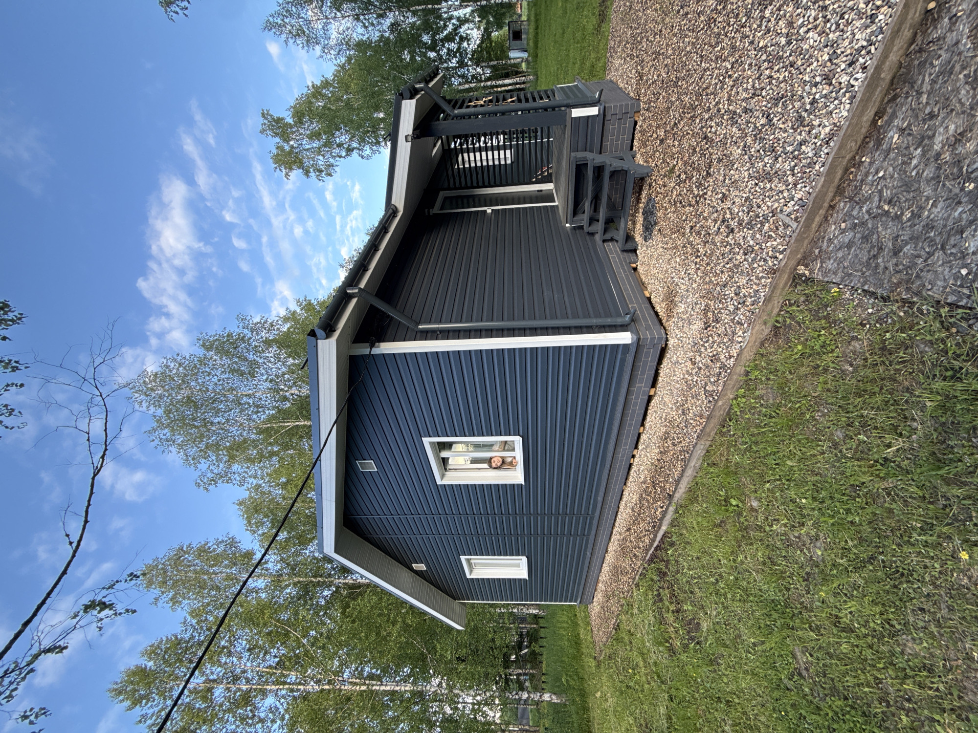 The image depicts a one-story, dark blue wooden cabin with white trim. The cabin has a porch with dark-stained wood steps and railings. The ground surrounding the cabin is covered with gravel, transitioning to grass. A person's face is visible in one of the windows. The cabin is situated in an outdoor area with trees and a partly cloudy sky. The location is Buzhaninovo, Russia.