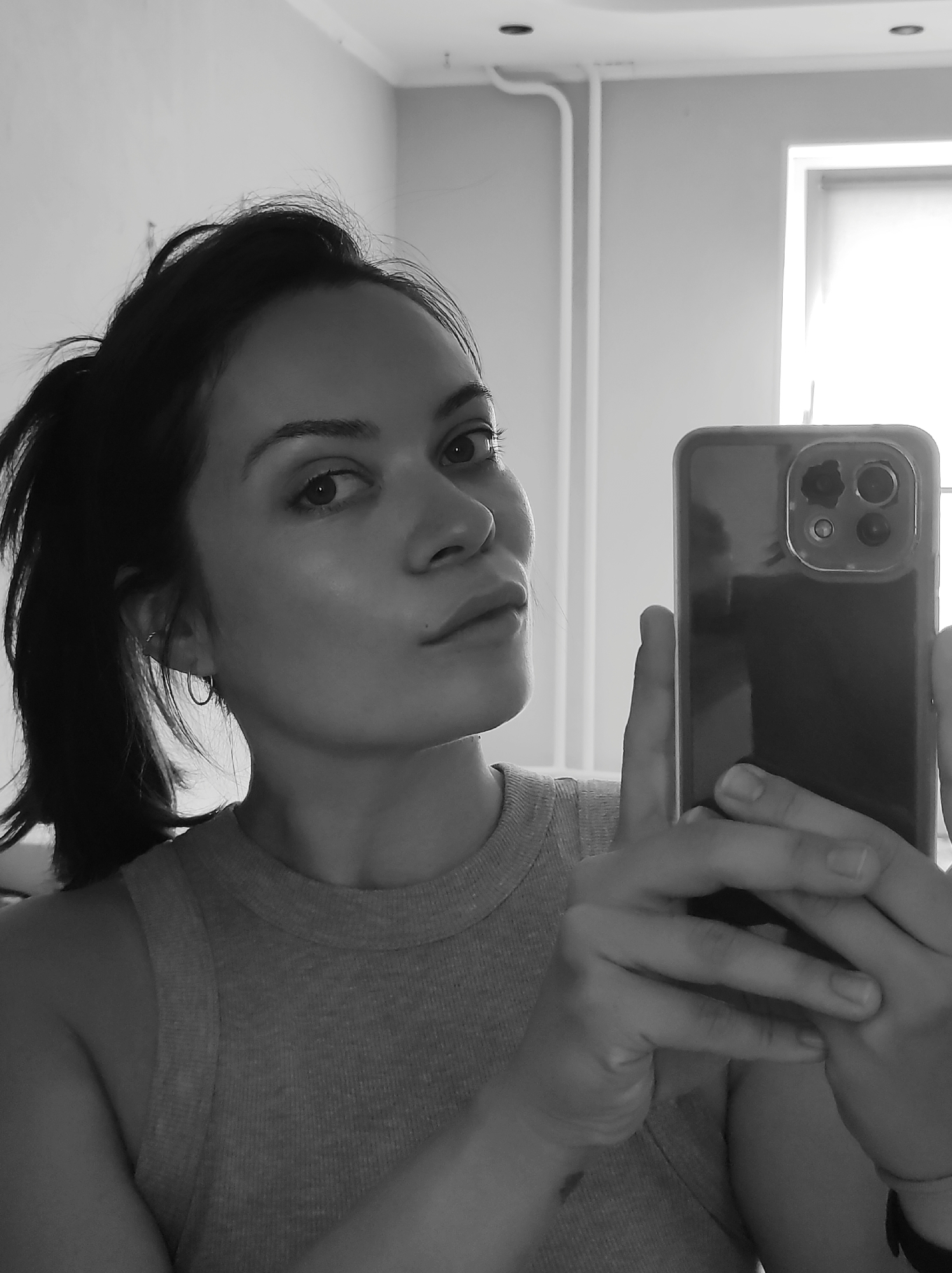 A black and white medium shot shows a woman taking a selfie in a room. She is looking towards the camera with her head slightly tilted. The woman has dark hair pulled back in a ponytail and is wearing a tank top. She is holding a smartphone in her right hand, taking the picture. The background of the image shows a plain wall with some electrical conduits and a window. The lighting suggests it is daytime. The scene appears to be an indoor residential setting, potentially an apartment. There is no specific identifying information within the image to pinpoint its location in Chaikovsky, Russia.