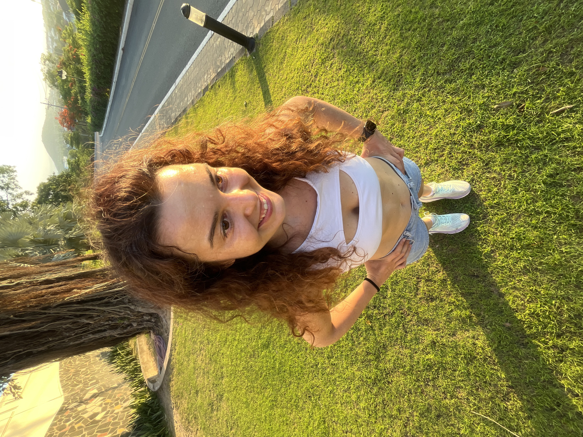 In Phu Thuy Ward, Vietnam, a young woman with curly auburn hair is captured in a high-angle shot. She is standing on a lush green lawn, bathed in the warm glow of the setting sun. Her white crop top with a cutout reveals her midriff, and she is wearing denim shorts and light blue athletic shoes. Her hands are placed on her hips, and she is smiling. Behind her, a large tree with exposed roots stands prominently, and a mosaic wall is visible to the left. The background features a paved walkway, a road curving uphill, and lush vegetation, including palm trees and flowering bushes, against a backdrop of rolling hills. A dark lamppost stands near the road.