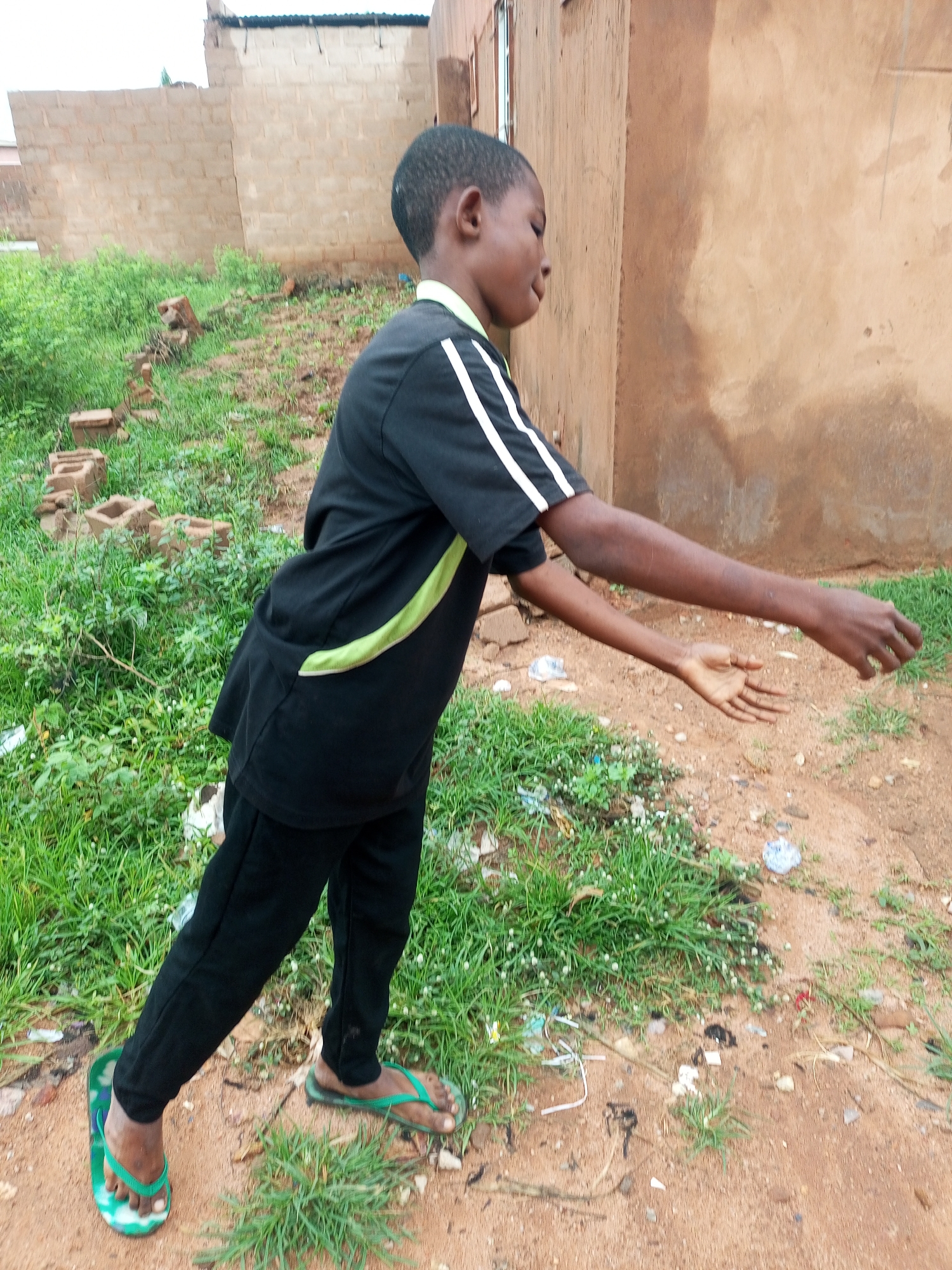 In an outdoor setting in an unknown city, a young boy of African descent stands in profile, facing right. He is wearing a black t-shirt with a lime green and white stripe across the chest and black pants. He is also wearing green flip-flops. The boy's right arm is extended outwards, with his hand open, as if reaching for something or in mid-gesture. His left arm is bent at the elbow and held closer to his body. The background consists of a dirt path with patches of green grass, small white wildflowers, and some scattered debris, including plastic bottles and paper. Behind the boy and to the right is a textured, brown, unfinished building wall. Further in the background, to the left, is another building with a light-colored brick wall. The overall scene appears to be an informal, possibly undeveloped, urban or semi-urban environment.