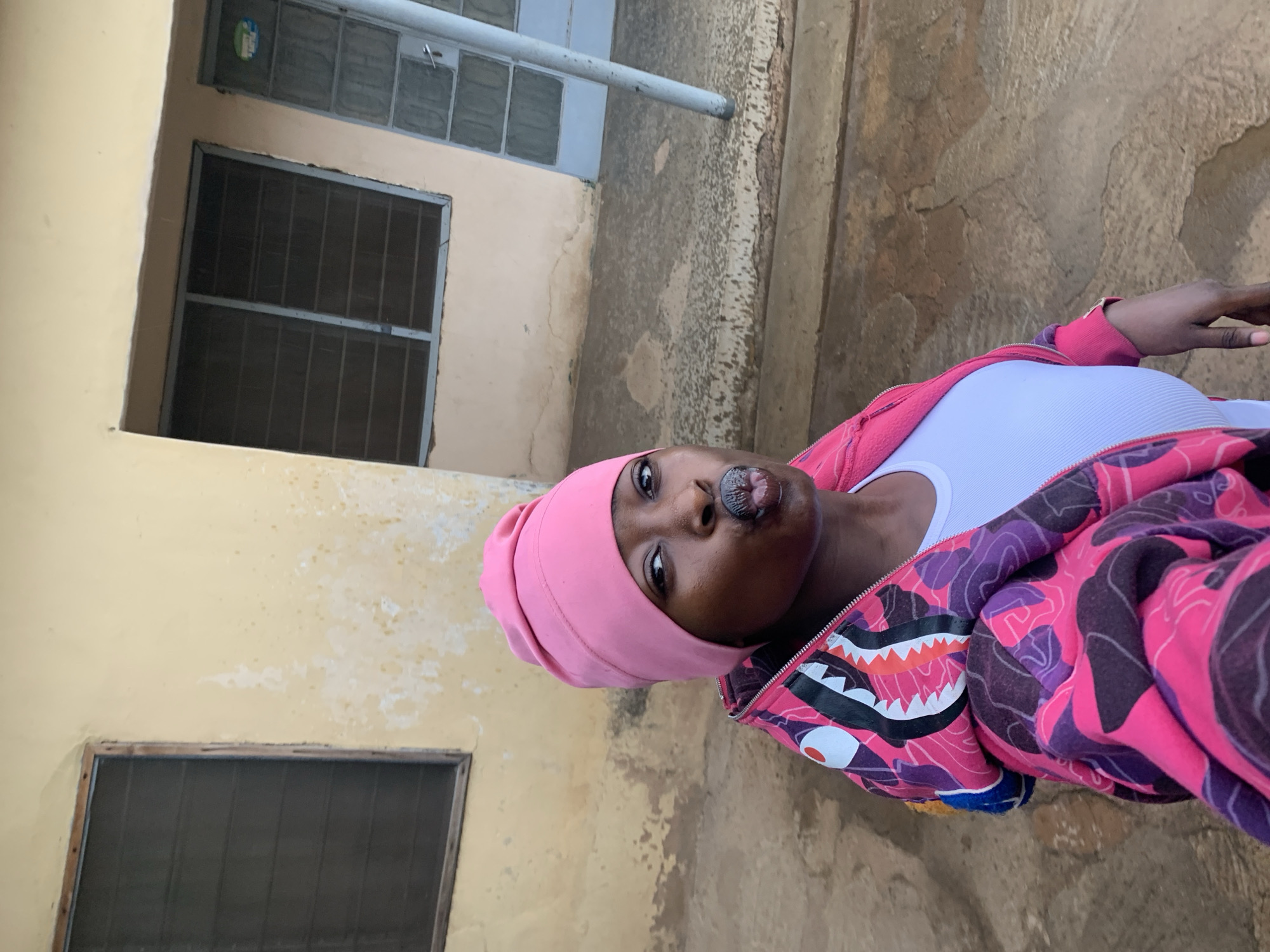 The image shows a person with dark skin wearing a pink head covering and a pink and purple patterned jacket with a shark-like design around the hood. They are also wearing a white top. The person is facing the camera with their lips pursed. The background consists of a yellowish building with windows fitted with metal bars and a concrete ground. The location is presumed to be Rafin Par, Nigeria, based on the user's request.
