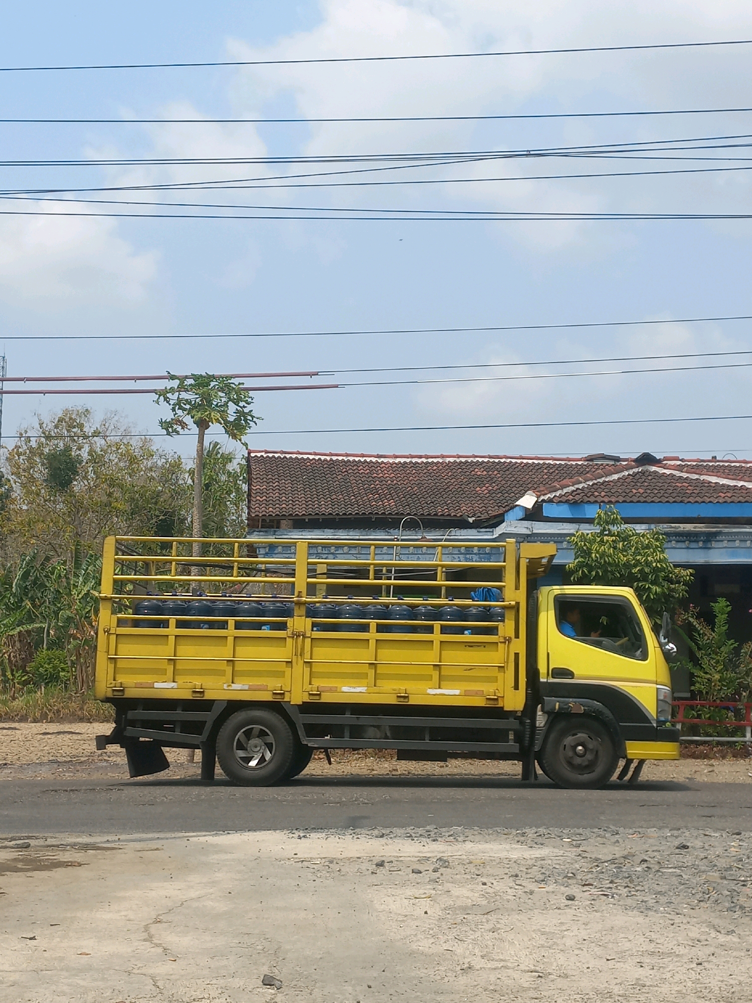 A yellow flatbed truck with a caged back is driving on a road in Baturetno, Indonesia. The truck is loaded with black barrels. The driver, wearing a blue shirt, is visible in the cab. The road is paved with some cracks and patches. In the background, there are houses with red tiled roofs, trees, and utility wires spanning across the sky. The sky is partly cloudy.