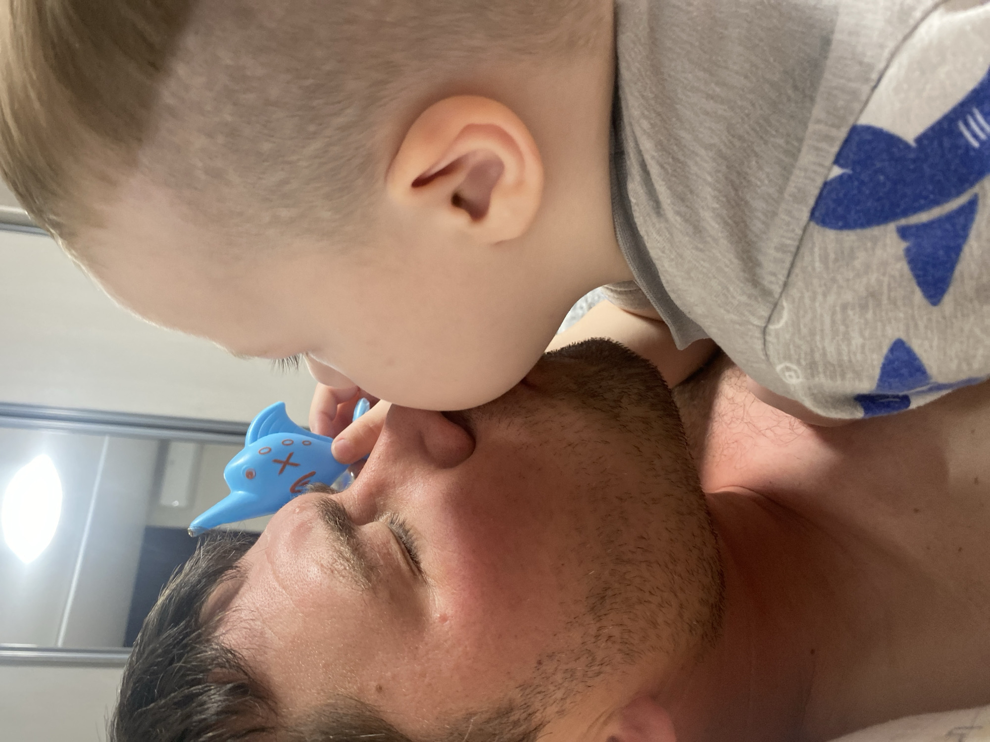 An overhead shot captures a tender and playful moment between an adult man and a young child indoors in Sochi, Russia. The man is lying on his back, eyes closed, with a close-cropped beard and dark hair. The child, with light blonde hair that is buzzed on the sides, is leaning over him, face-to-face, almost touching noses. The child wears a light grey t-shirt adorned with blue shark illustrations, one of which is clearly visible on the shoulder.
In a whimsical touch, the child holds a vibrant blue, fish-shaped toy with red and white markings (eyes, an "X" for a mouth, and stripes) just above the man's nose. The man's closed eyes suggest he is either resting, enjoying the interaction, or playfully engaging with the child. The scene conveys strong emotions of affection, warmth, and joy between the two individuals.
The background shows a bright, reflective surface, possibly a mirror, with a light fixture casting a glow, indicating an indoor setting like a bedroom or bathroom. No specific location cues for Sochi, Russia are visible within the image itself. There is no visible text. The time of day cannot be determined, and weather conditions are not visible as the scene is indoors.