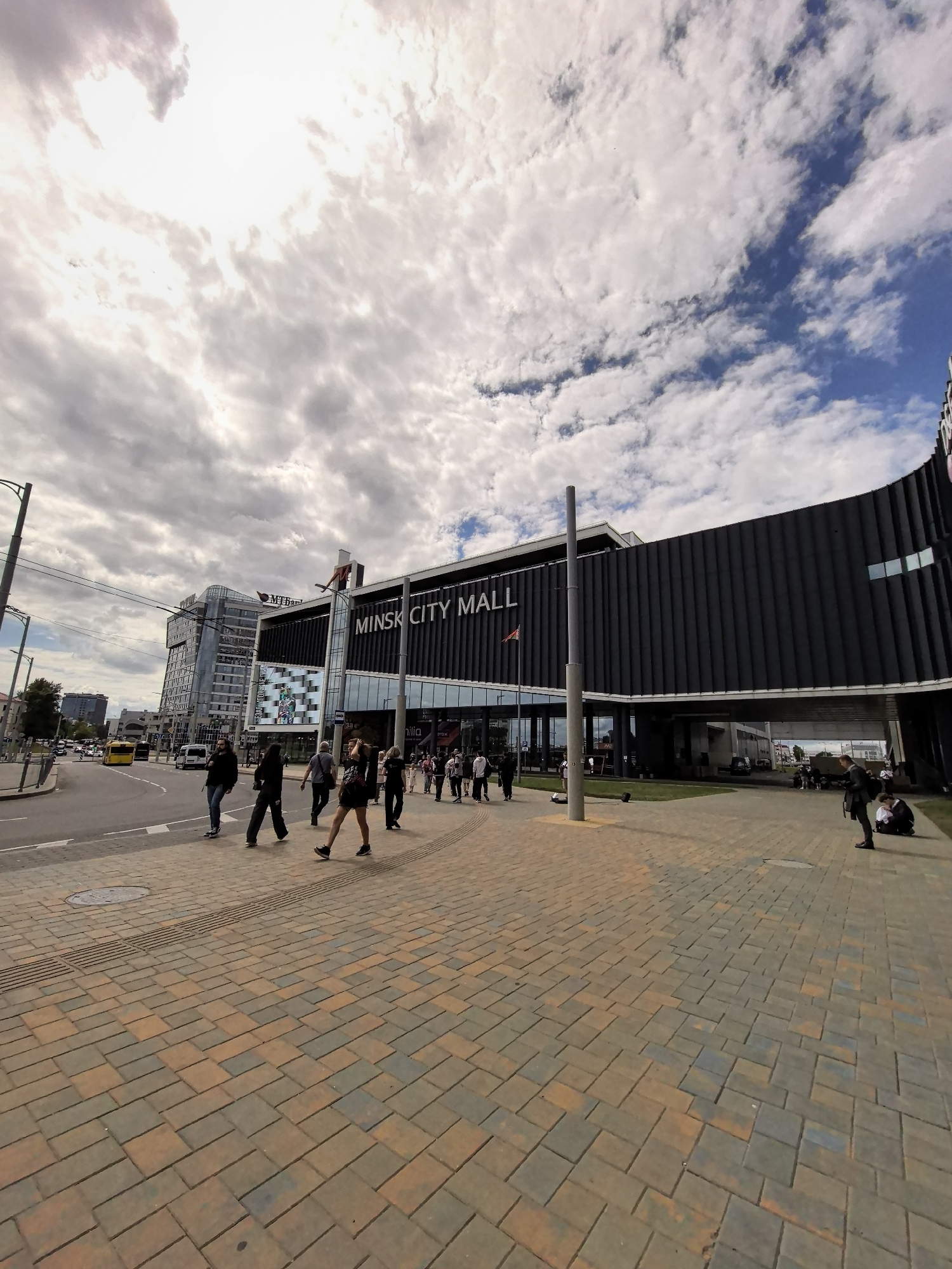 A wide-angle, low-angle shot captures the facade of Minsk City Mall in an urban setting under a partly cloudy sky. The mall's modern, dark-paneled exterior curves along the street, with a prominent sign displaying "MINSK CITY MALL" in white lettering. A pedestrian walkway paved with multicolored rectangular stones leads towards the mall. Several individuals are visible, some walking across the street, others on the walkway. In the background, a multi-story glass-fronted building stands, and traffic, including a yellow bus, moves along the road. The scene is illuminated by daylight, with the sun partially obscured by clouds.
