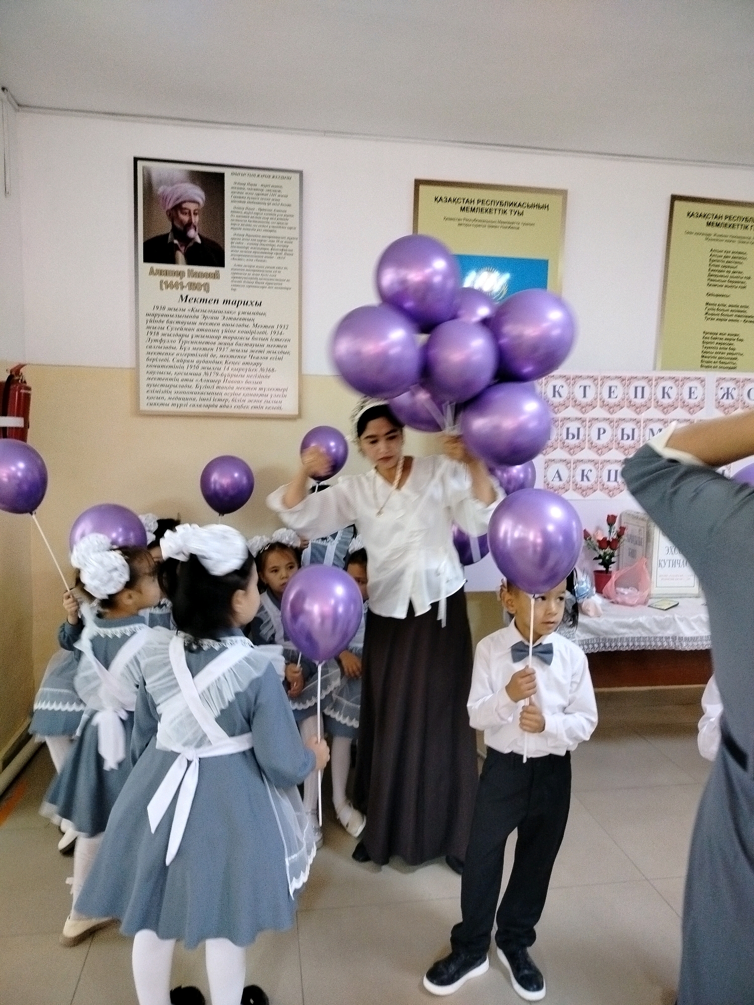 A group of children, mostly girls in grey dresses with white pinafores and bows, and one boy in a white shirt and black pants with a bowtie, are gathered indoors. They are holding numerous purple balloons. A woman in a white blouse and dark skirt stands behind some of the children, holding a large cluster of balloons. Behind the group, posters are mounted on the wall, one featuring a portrait and text, another with the Kazakh text "ҚАЗАҚСТАН РЕСПУБЛИКАСЫНЫҢ МЕМЛЕКЕТТІК ТУЫ". The environment appears to be a school or community hall, with a tiled floor and a beige wall. The city of Көлкент, Kazakhstan is mentioned as the location.