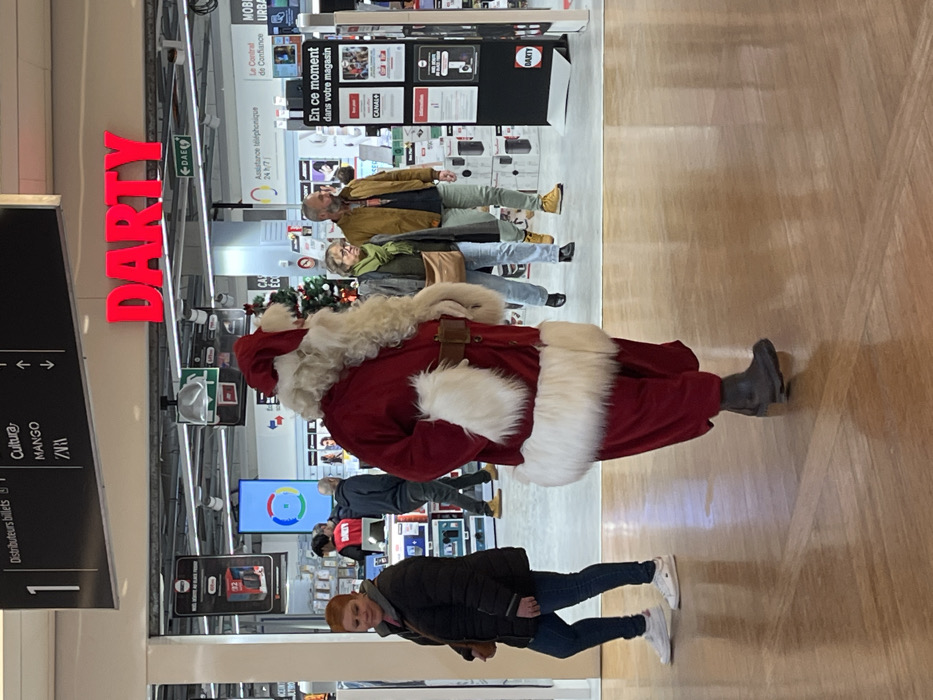 The scene is set within the concourse of a shopping mall, identified as the Centre Commercial Dijon Toison d'Or in Dijon, France. The highly polished, light brown wooden floor reflects the overhead lighting and the figures present.
The primary subject is an individual dressed in a complete Santa Claus costume, comprising a red velvet suit with white fur trim on the collar, cuffs, and hem, a matching red hat, and a long white beard. This figure wears dark boots and walks from right to left across the foreground, with hands resting in the costume's pockets.
To the left, a woman with short reddish-brown hair walks from left to right. She wears a dark quilted jacket, blue jeans, and white sneakers, carrying a light brown shoulder bag.
In the mid-ground, a couple walks from right to left. The woman, with light hair and glasses, wears a dark jacket, a light green scarf, and light blue jeans, carrying a brown shoulder bag. Alongside her, a man with a beard and short dark hair wears a brown jacket, a dark t-shirt, light green trousers, and tan boots.
The background reveals the entrance to a DARTY electronics store, prominently marked with a large red illuminated sign. Inside DARTY, various electronic products are displayed, alongside a large vertical digital screen showing a multi-colored circular logo, and a small, decorated Christmas tree. Overhead mall signage indicates the presence of "Cultura," "MANGO," and "ZARA," as well as "Distributeurs billets" (ATMs), and a "DAE" (Automated External Defibrillator) sign. A black information stand displaying "En ce moment dans votre magasin" (Currently in your store) and "CANAL+" branding is situated to the right. Additional indistinct figures are visible further in the background, both inside and outside the store.