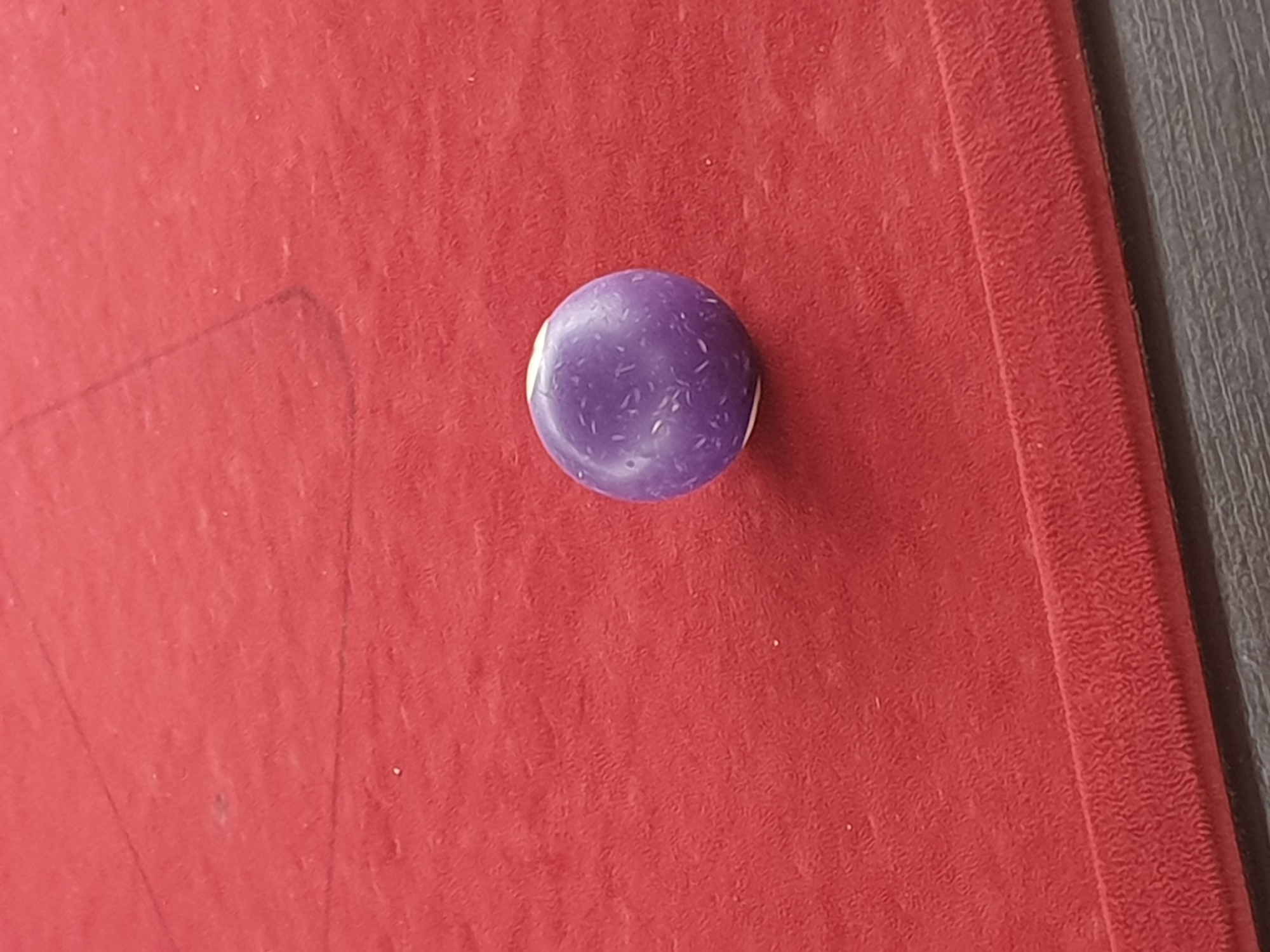 The image shows a close-up, slightly angled view of a purple billiard ball resting on a red billiard table. Faint, dark lines, likely part of the billiard table's markings, are visible on the surface, forming geometric shapes. The texture of the red felt is prominent, with a slight sheen indicating a smooth surface. The ball itself has a matte finish with small, lighter-colored speckles scattered across its surface. The bottom edge of the image reveals a dark, horizontal surface, possibly the side of the table or a shelf. There is no indication of the city of Lokoja, Nigeria, in this image; the scene is solely focused on the billiard ball and table.