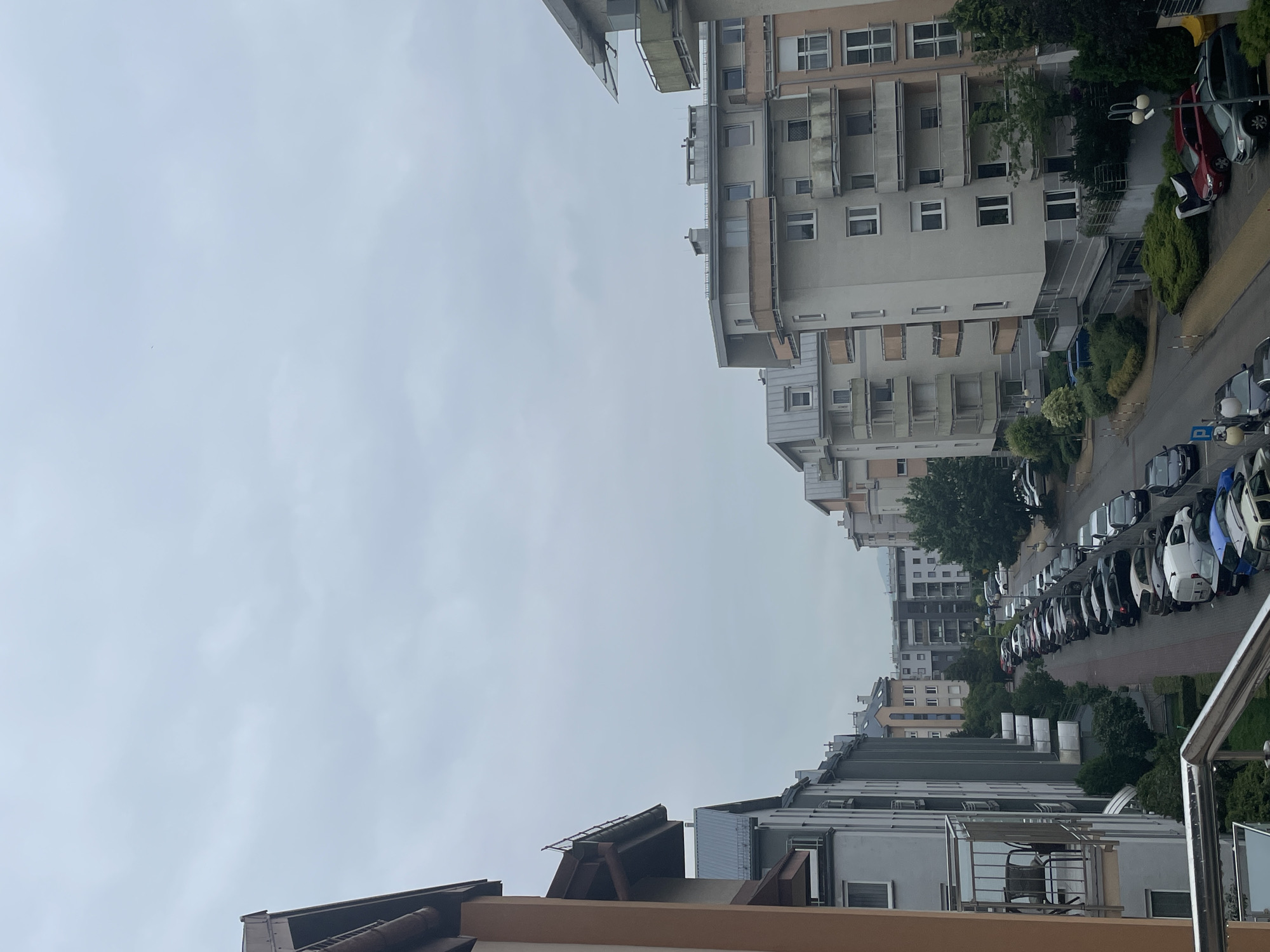 The image depicts a street scene in a residential area, likely an apartment complex, under an overcast sky. The perspective is from an elevated position, possibly a balcony or window, looking down a street lined with multi-story buildings on both sides. The buildings are modern, with balconies and numerous windows. On the left side of the street, a balcony with a chair is visible. On the right side, the buildings have a similar architectural style with beige and white facades. The street is filled with parked cars of various colors, including black, silver, white, blue, and red. Some of the cars are parked along the curb, while others are in designated parking spaces. There are trees and green spaces interspersed between the buildings and along the street. The overall impression is of a typical urban residential environment.