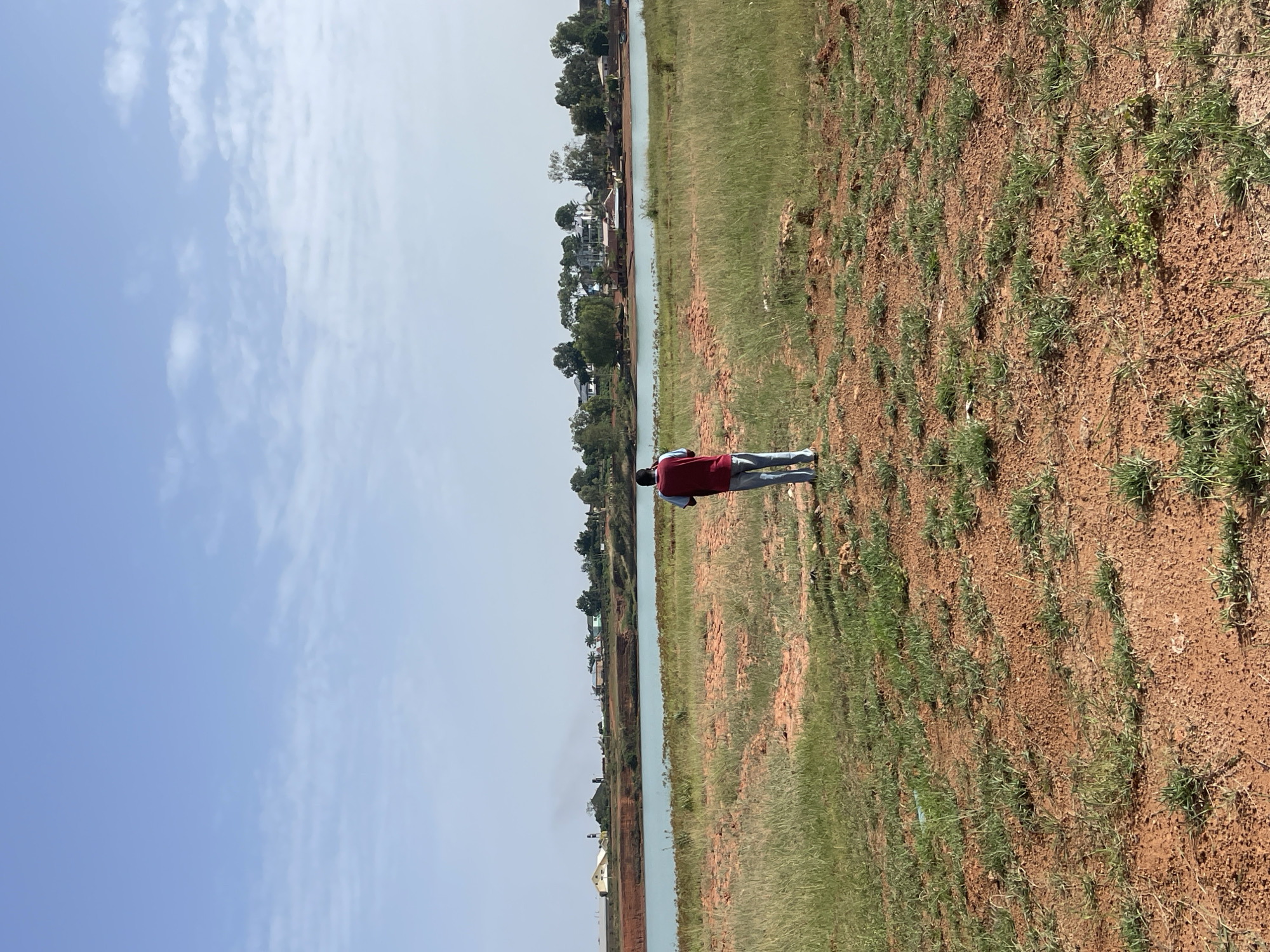 A person wearing a red shirt and blue pants stands on a plot of land in an unspecified location in Jos, Nigeria. The land comprises reddish soil with sparse grass cover. A body of water, possibly a river or lake, runs parallel to the plot. Trees and buildings can be seen on the far side of the water. The sky above is blue with scattered clouds.