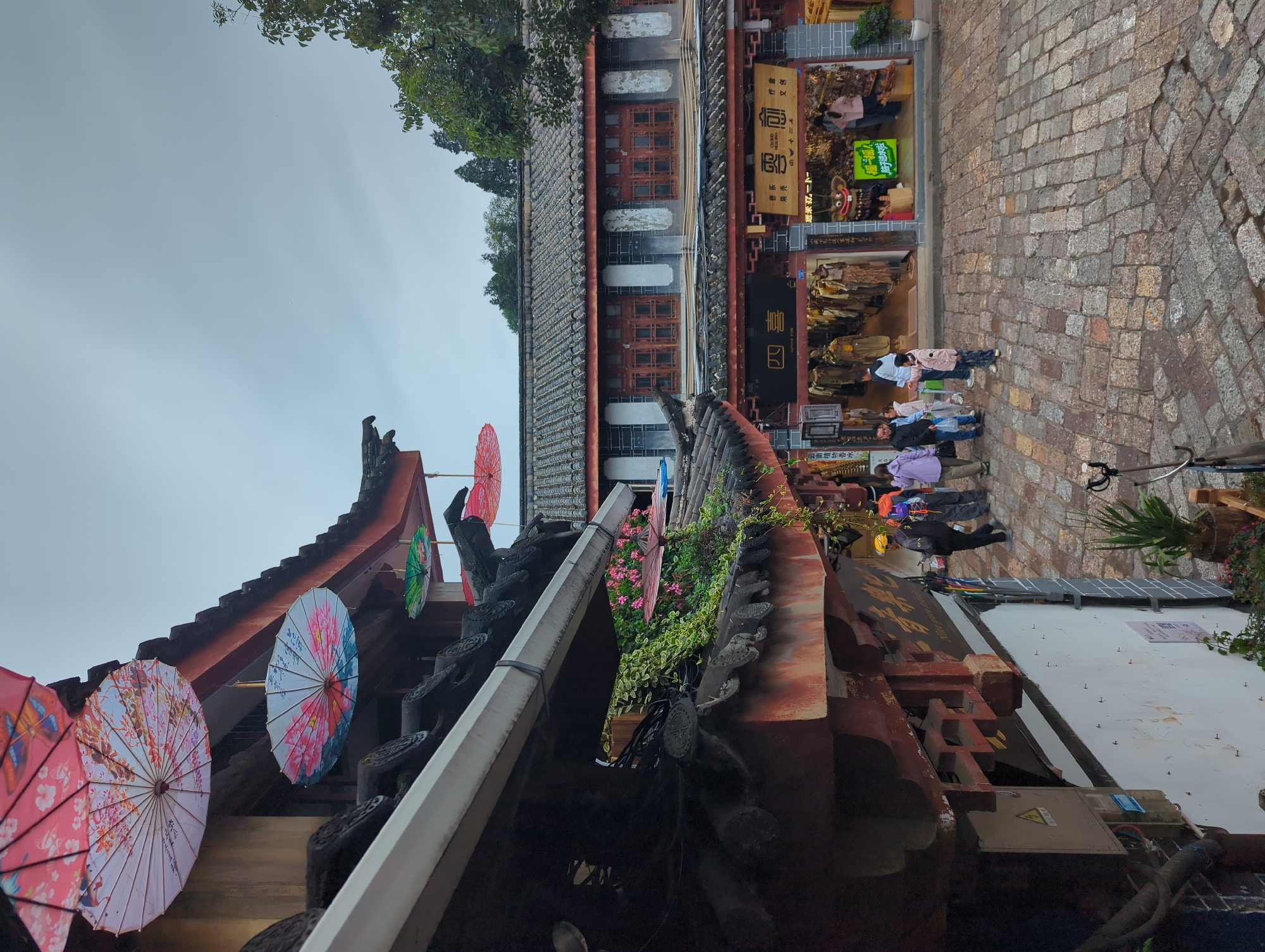 In Gucheng District, China, an overcast sky looms over a street scene featuring traditional Chinese architecture. The foreground is dominated by the angled roofline of a building adorned with ornate eaves and multiple decorative paper umbrellas. These umbrellas, in vibrant pinks, whites, and greens, are suspended by strings, adding a splash of color to the scene. Green vines with small pink flowers cascade down the side of the building.
The street itself is paved with cobblestones, showing signs of moisture. Several individuals are visible walking and interacting on the street. In the mid-ground, a row of shops with traditional tiled roofs and wooden window frames line the street. One shop displays clothing on mannequins, while another features a display with a cartoonish, grinning character. People can be seen entering and browsing within these establishments. The overall atmosphere suggests a bustling market or shopping area in a historic district.