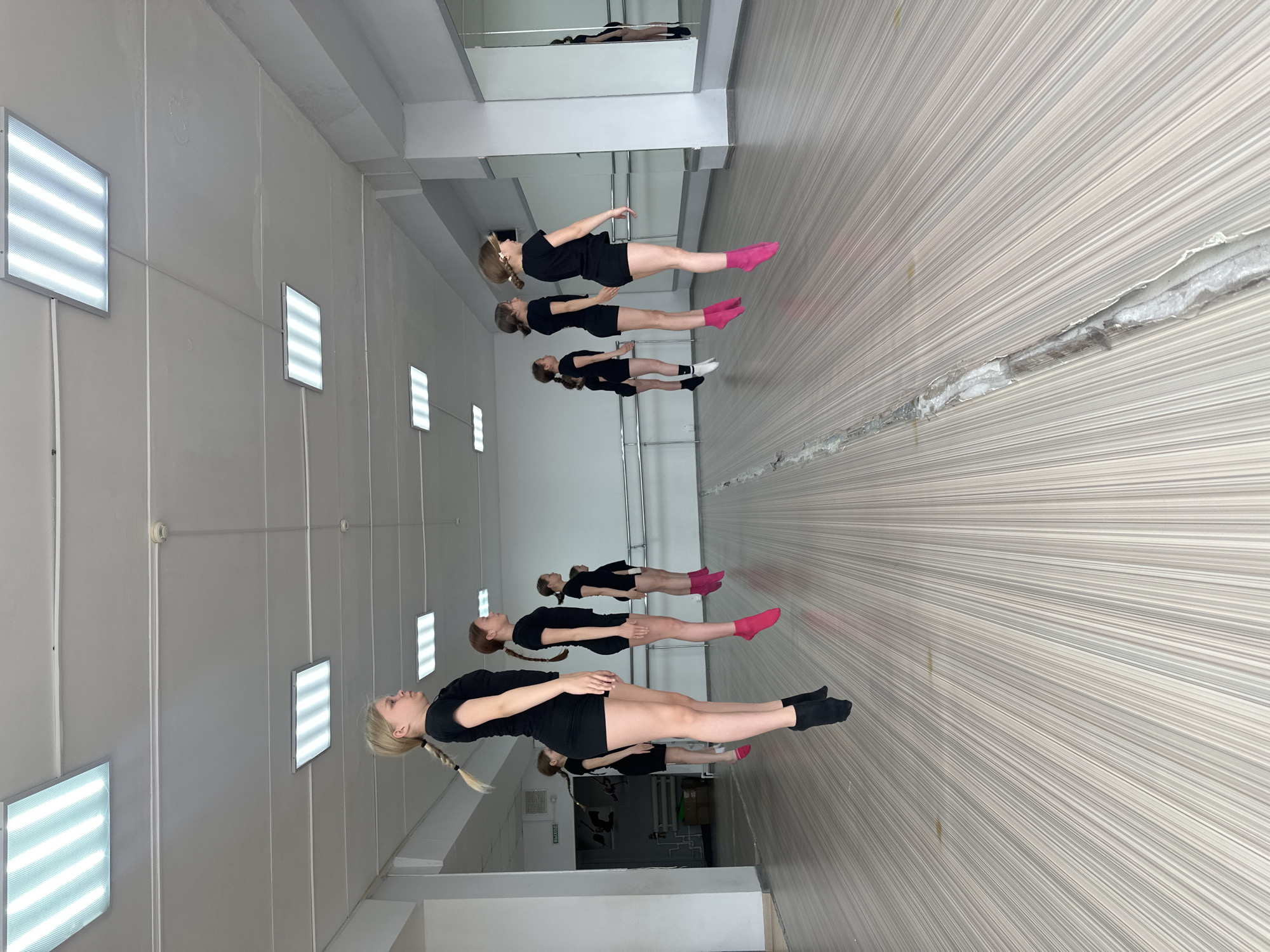 A dance class is taking place in Votkinsk, Russia, inside a studio or gymnasium. Six girls are wearing black dance outfits and practicing their poses. The foreground of the room has a reflective floor. The room is lit by rectangular overhead lights.