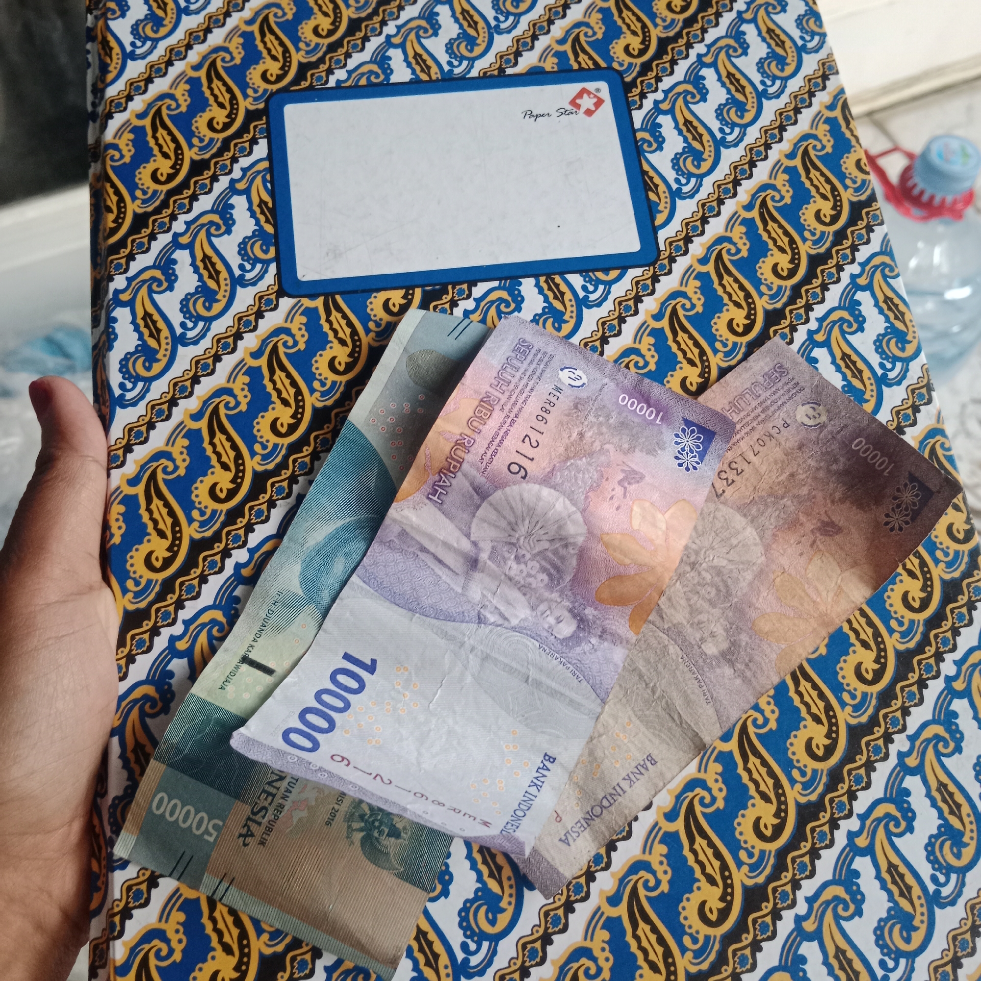The image shows a hand holding several Indonesian Rupiah banknotes on top of a patterned book cover. The book cover features a blue, yellow, and black paisley-like pattern. The banknotes visible are denominations of 50,000 and 10,000 Rupiah. The 50,000 Rupiah note is green and brown, while the 10,000 Rupiah notes are purple and brown. A blue label with white text and a small red and white logo is present on the book cover, suggesting it might be a notebook or binder. In the background, a blurry plastic water bottle with a blue cap and a red tie is visible. The scene appears to be indoors, possibly in a home or office setting. There is no specific information or visual cues in the image that indicate the location within Tegowanu Kulon, Indonesia.